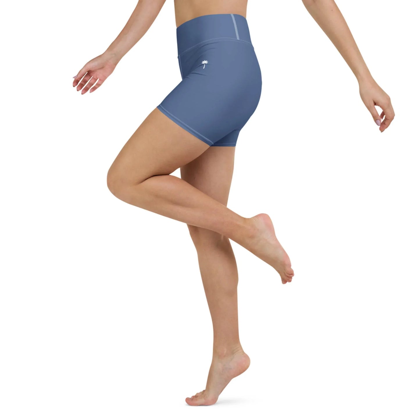 A woman models Printfuls Brickell Yoga Shorts for women in blue, featuring a high-waisted design. Posing on one leg with arms lifted, only her torso and legs are shown against a white background.
