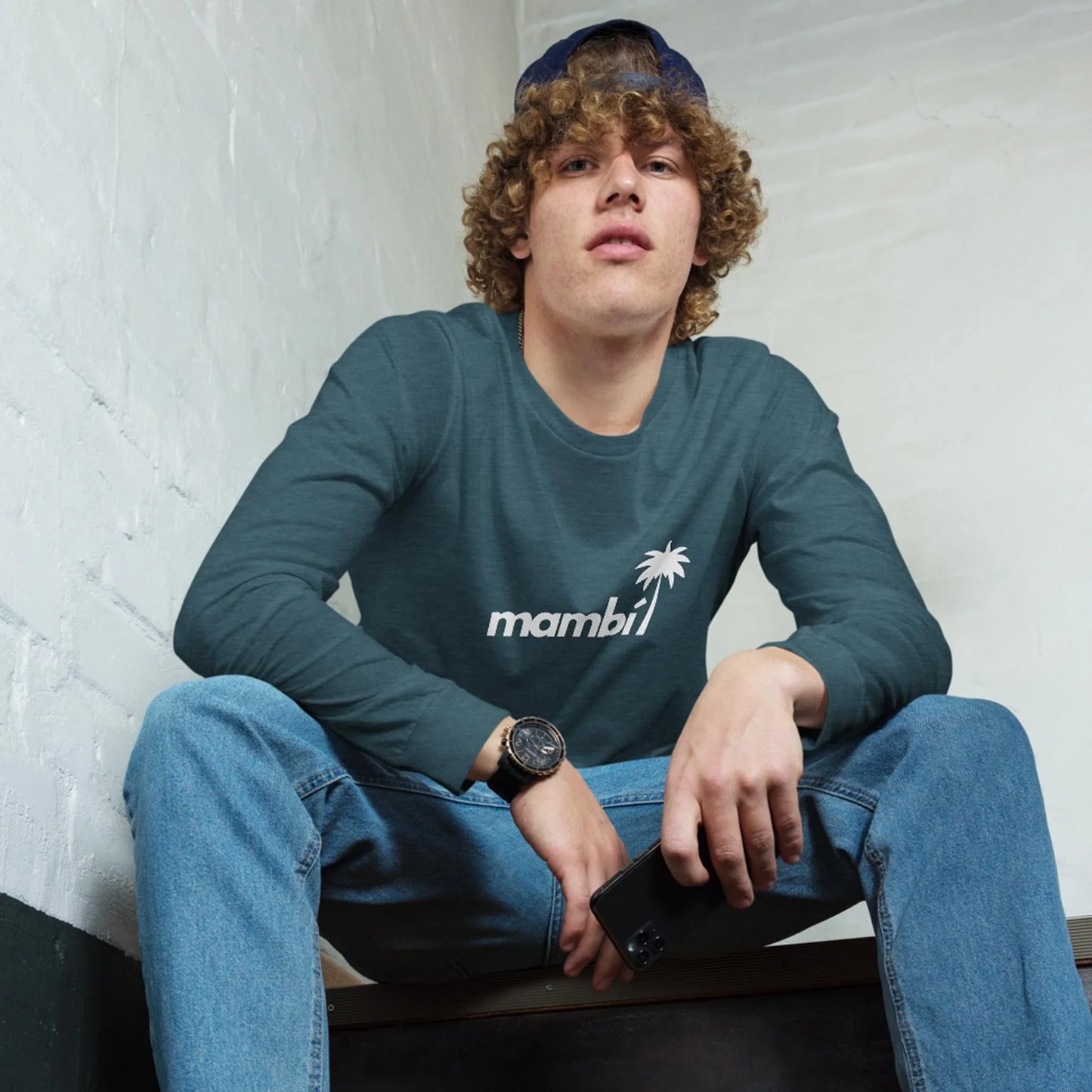 A young man with curly hair, wearing a blue cap and a teal long-sleeve Mambí shirt with a palm tree logo, blue jeans, and a watch, sits on stairs holding his smartphone, looking up thoughtfully.