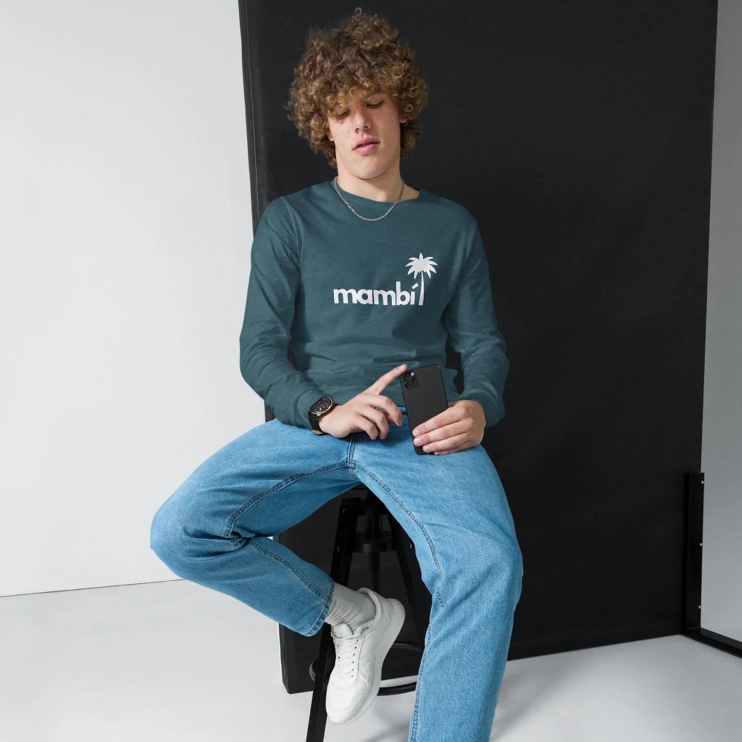 A young person with curly hair sits on a stool against a black and white background, wearing a blue Mambí long-sleeve shirt, jeans, white sneakers, and holding a smartphone.