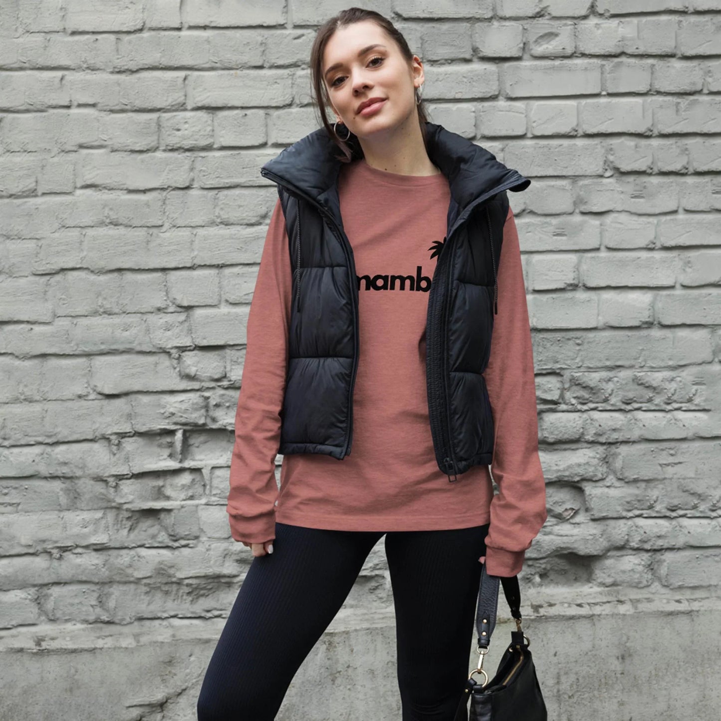 A woman stands in front of a gray brick wall, wearing a pink long-sleeve Mambí shirt with black text, a black puffer vest, black pants, and holding a black handbag. She has her hair tied back and is looking at the camera.
