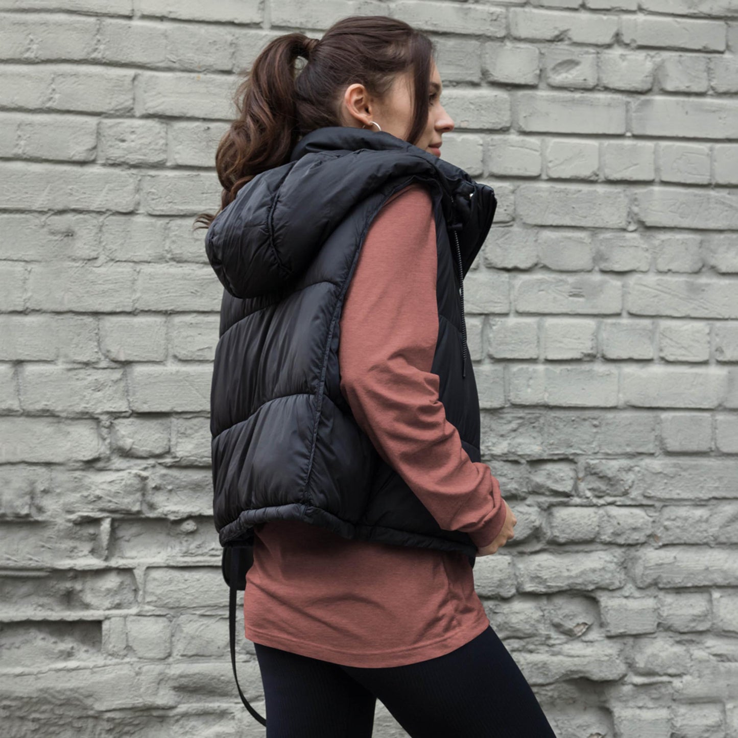 A woman with a ponytail, wearing a Mambí black puffer vest over a long-sleeved mauve shirt and black pants, stands sideways against a light gray brick wall.