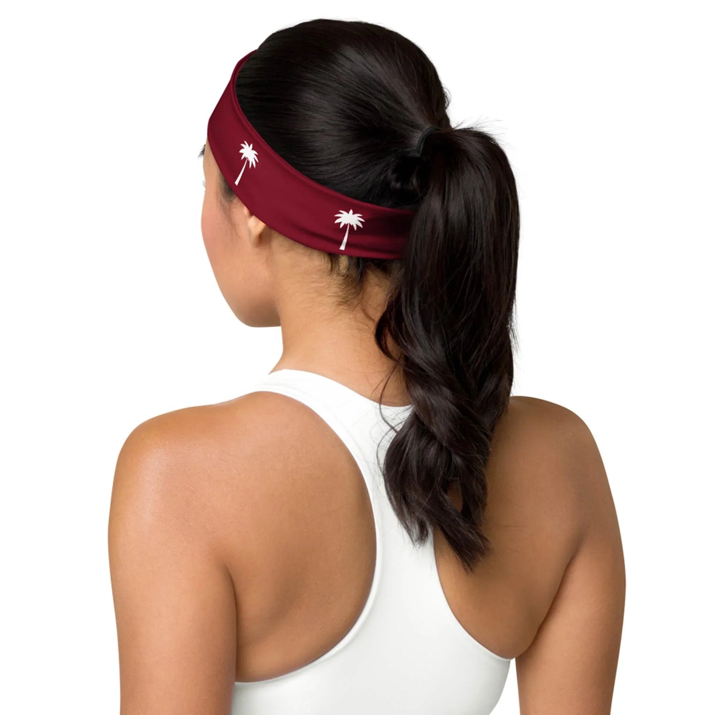 A woman with dark hair in a ponytail, wearing a white racerback top and a maroon Mambí headband with white palm tree designs, is shown from behind against a plain background.