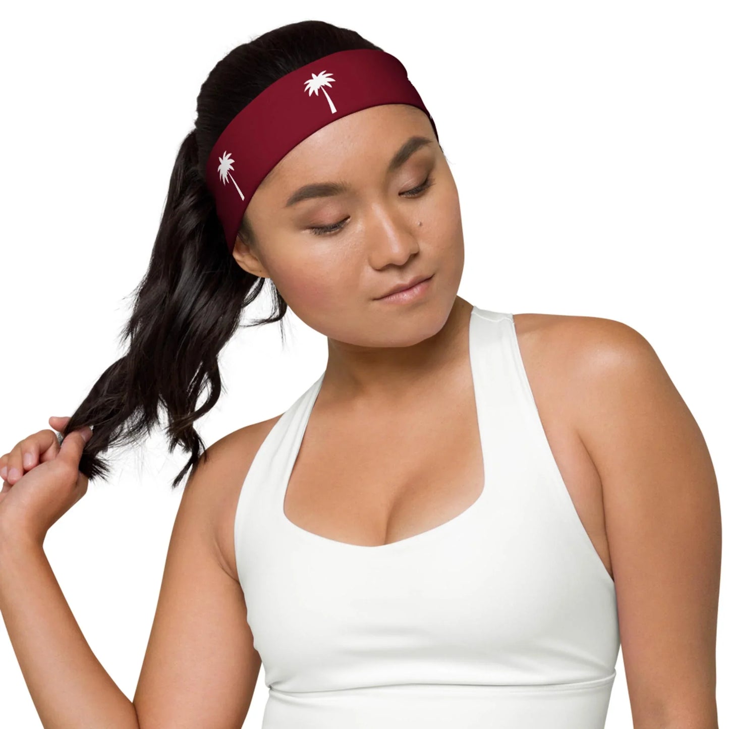 A woman in a white sports top looks down while holding her dark ponytail. She wears a maroon Mambí headband decorated with small white palm tree designs.