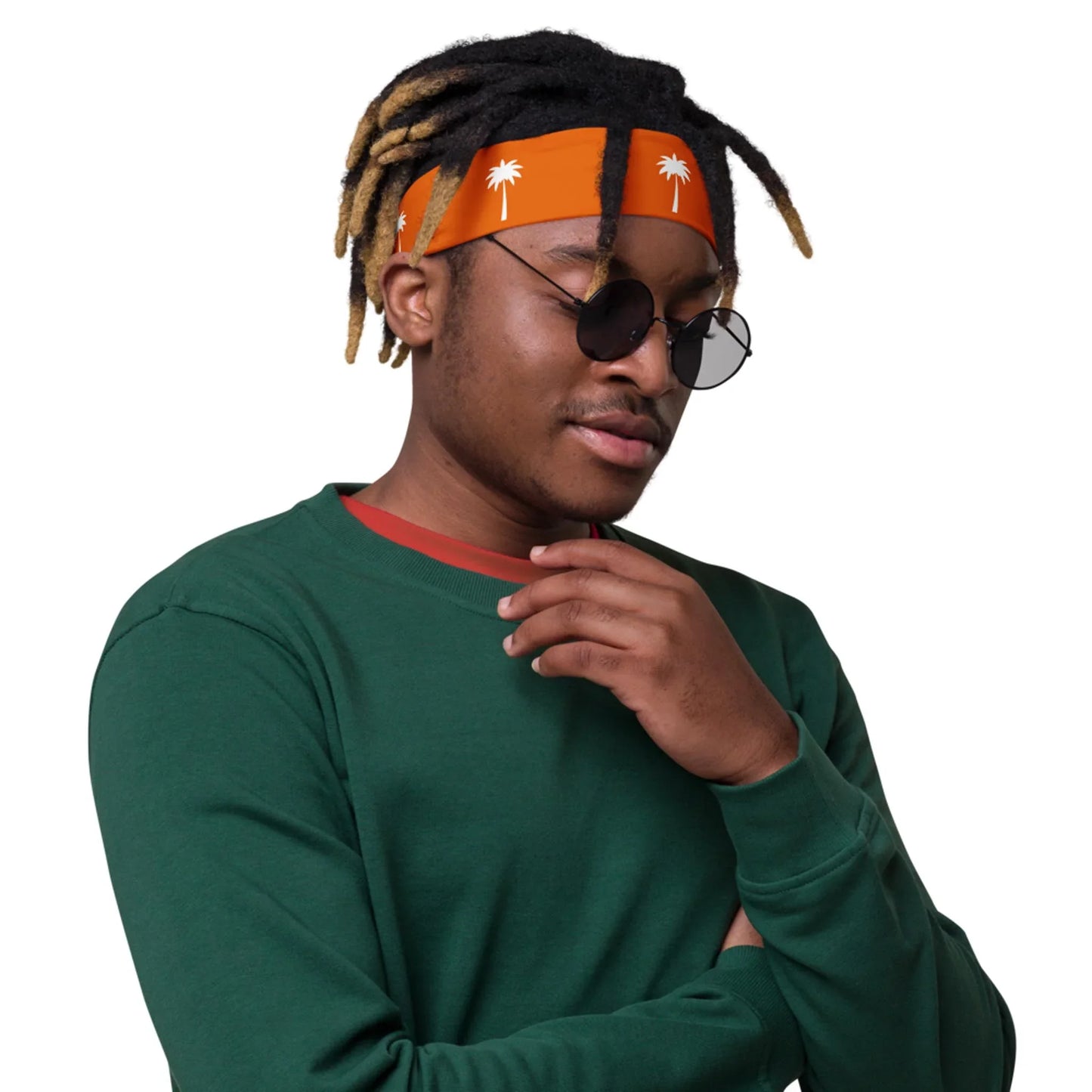 A person wearing round black sunglasses, a Mambí orange headband with white palm trees, and a green sweatshirt stands with one hand touching their chin against a white background.