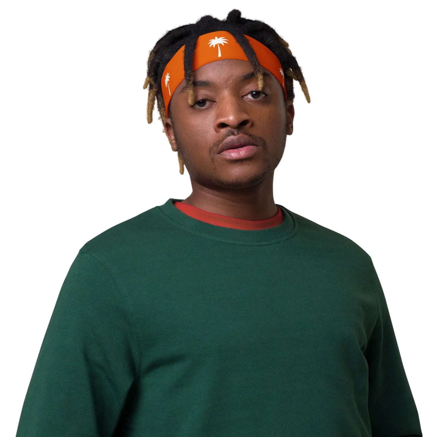 A man with short dreadlocks and an orange headband featuring white palm tree designs wears a dark green Mambí sweatshirt over a red shirt, standing against a plain white background.