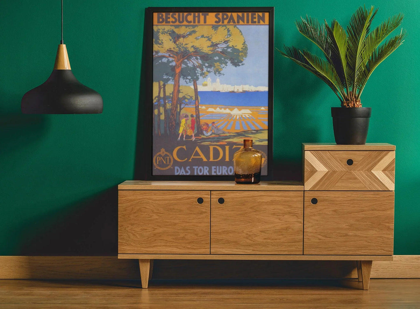 A modern wooden sideboard sits against a green wall, topped with Printfuls Cadiz Reprinted Vintage Poster on premium luster paper (24x36), a brown glass bottle, and a potted palm. A black pendant lamp hangs above, finishing the look.