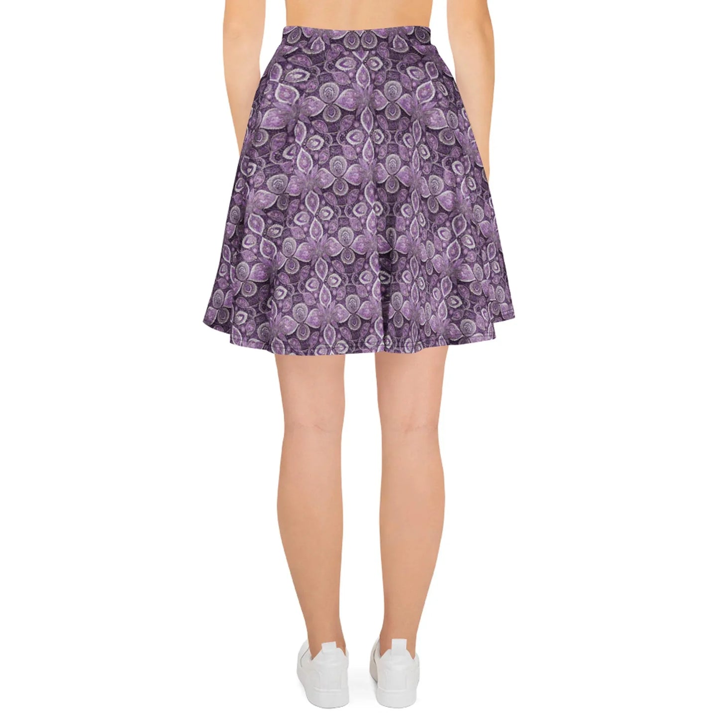 A woman faces away, wearing the Printful Candelaria Skater Skirt for women—a purple patterned skirt with pleats and an elastic waistband—showing a flattering silhouette. She pairs it with white sneakers.