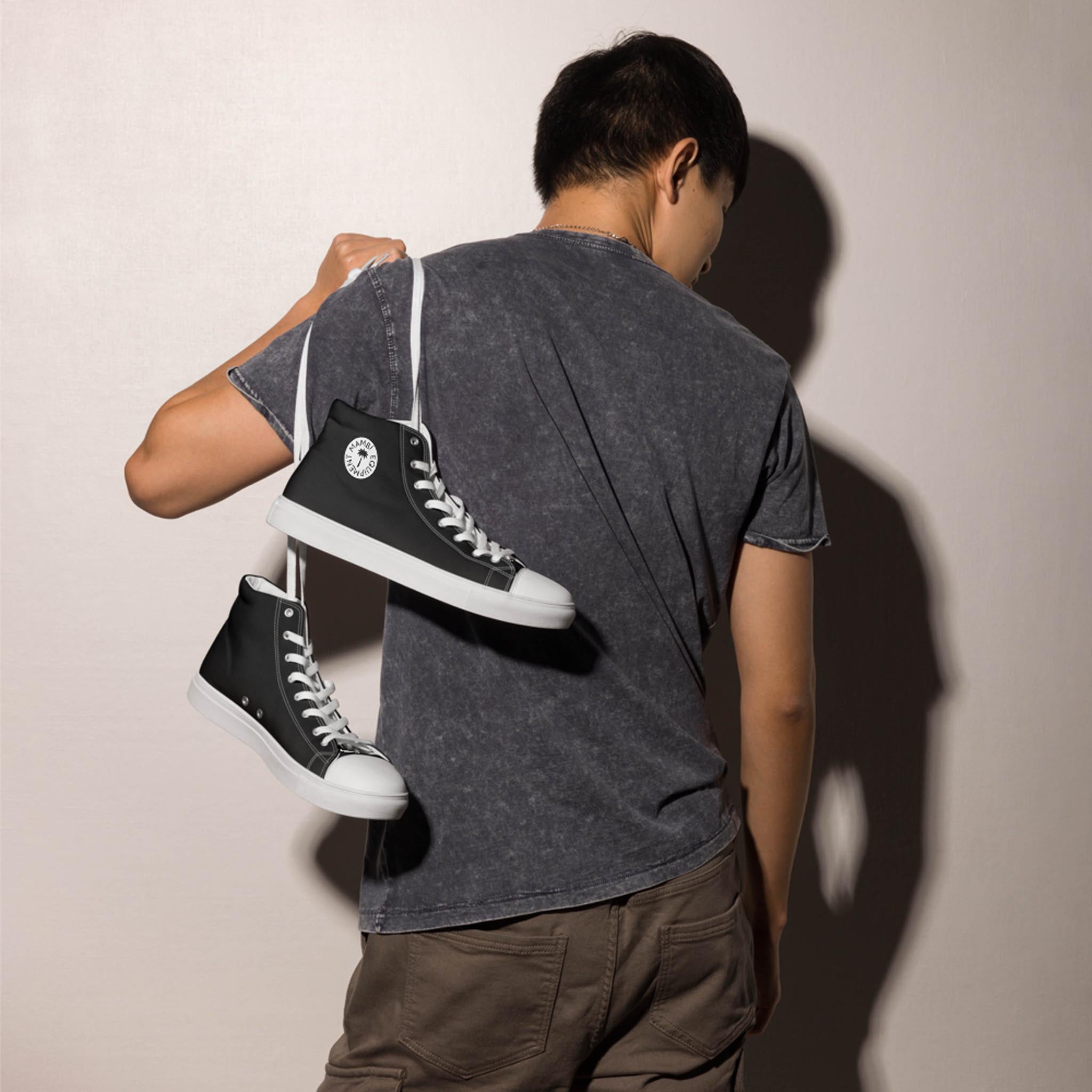 A person stands with their back to the camera, holding a pair of black high-top sneakers slung over their shoulder by the laces. They wear a dark gray t-shirt and brown pants, casting a shadow on the wall.