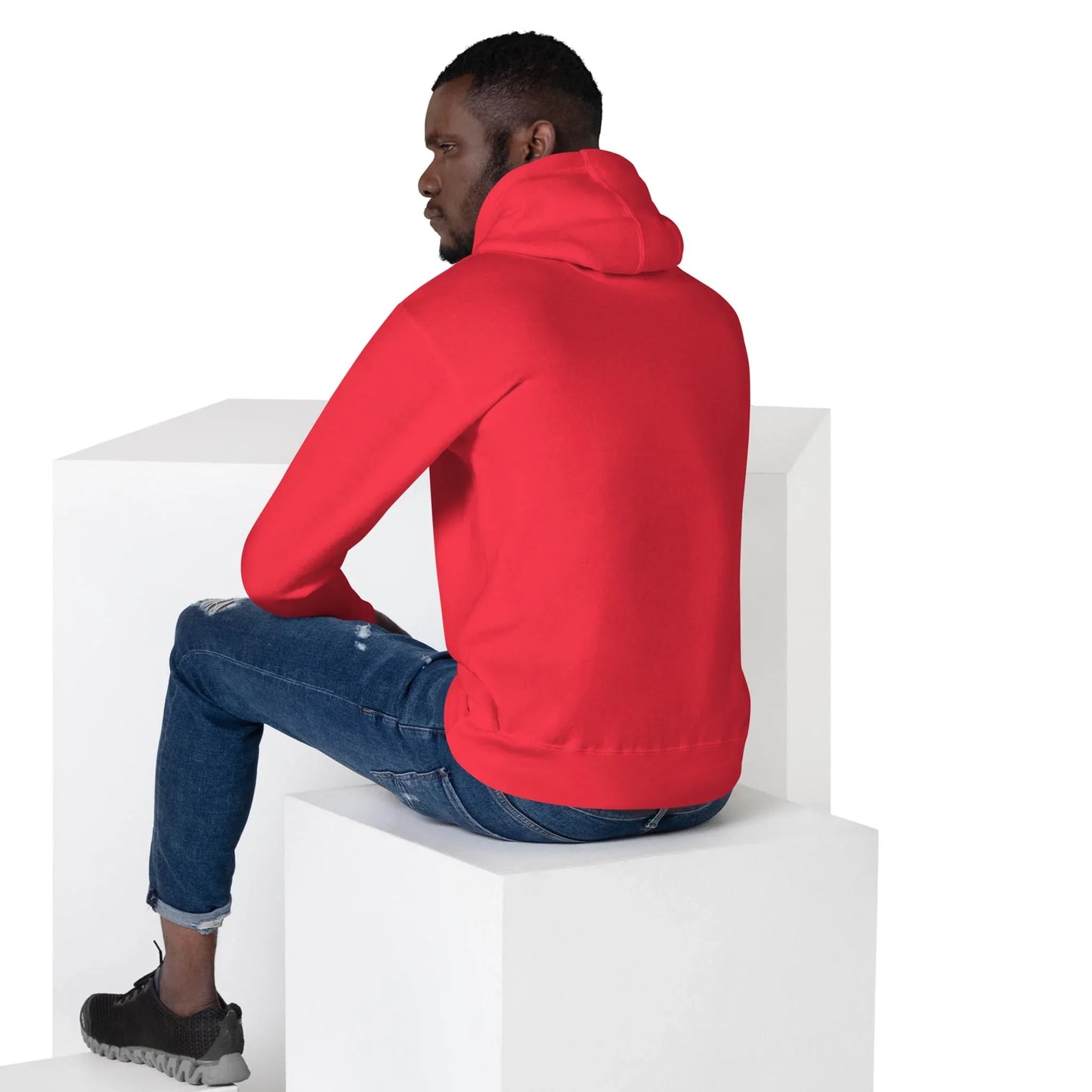 A man wearing a bright red Mambí hoodie and ripped blue jeans sits on a white block, facing away from the camera in a minimalist white studio setting.