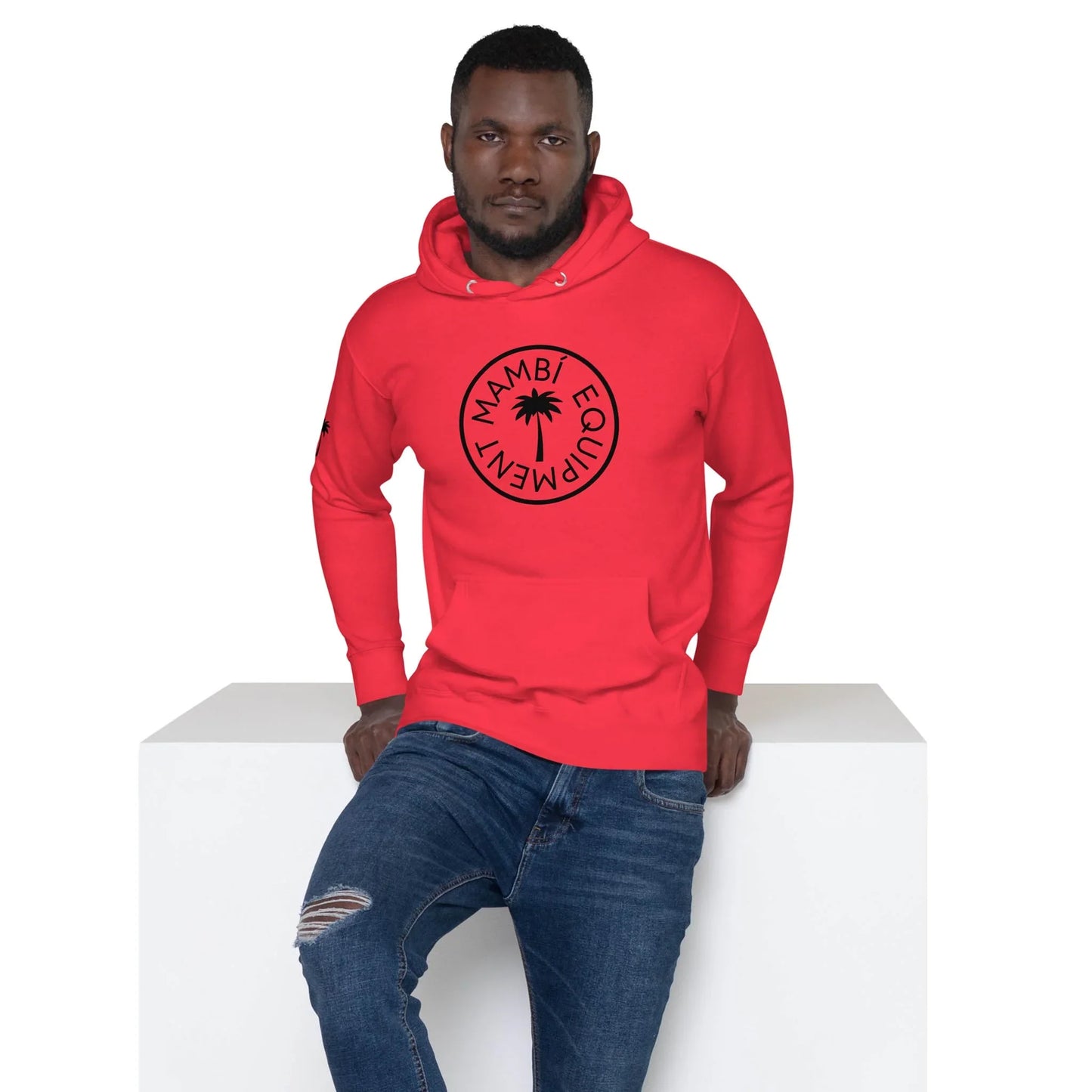 A man in a bright red hoodie featuring the circular black Mambí logo and ripped-knee jeans sits on a white rectangular block, looking directly at the camera.