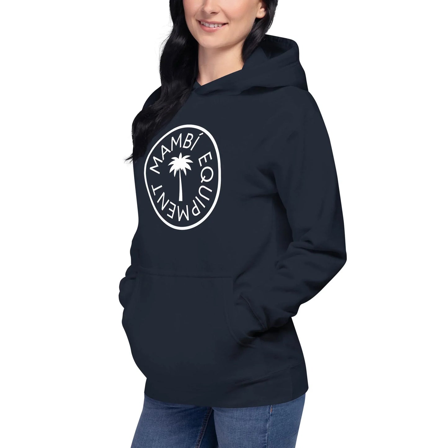 A woman in a black Mambí hoodie with a white circular logo and palm tree stands with her hands in the front pocket, smiling slightly. She is wearing blue jeans.