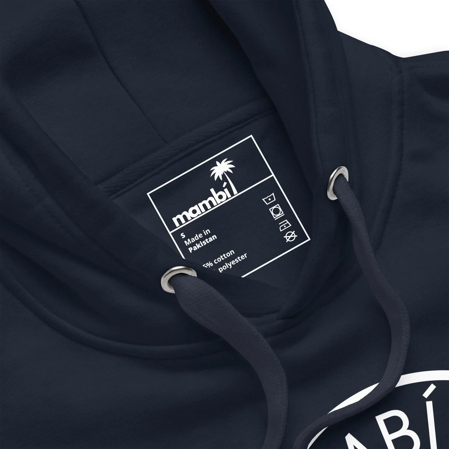 Close-up of a navy blue hoodie with a white label inside the collar showing the Mambí brand, a palm tree logo, care symbols, and text stating Made in Pakistan, 80% cotton, 20% polyester.