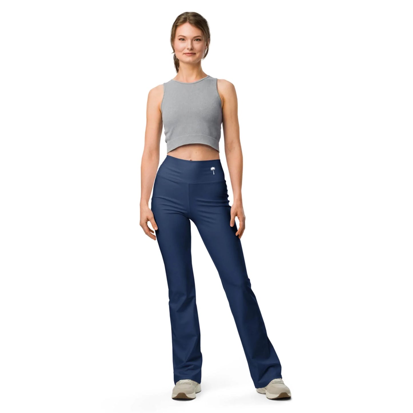 A woman poses casually in Printfuls Carolina Flared Performance Leggings for women, navy blue and high-waisted, paired with a gray sleeveless crop top and beige sneakers against a plain white background.