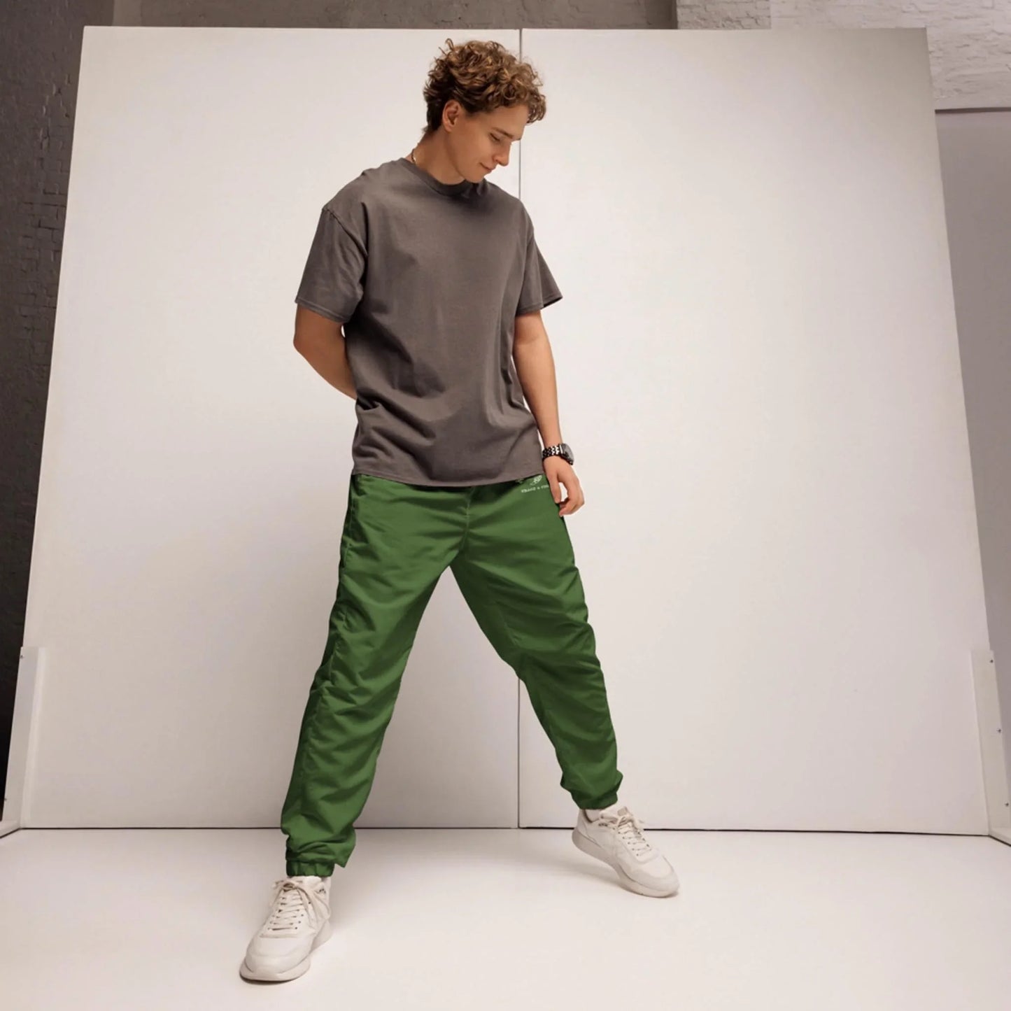 A person stands indoors against a white backdrop, wearing a gray Mambí t-shirt, green pants, and white sneakers, looking down with hands in pockets.