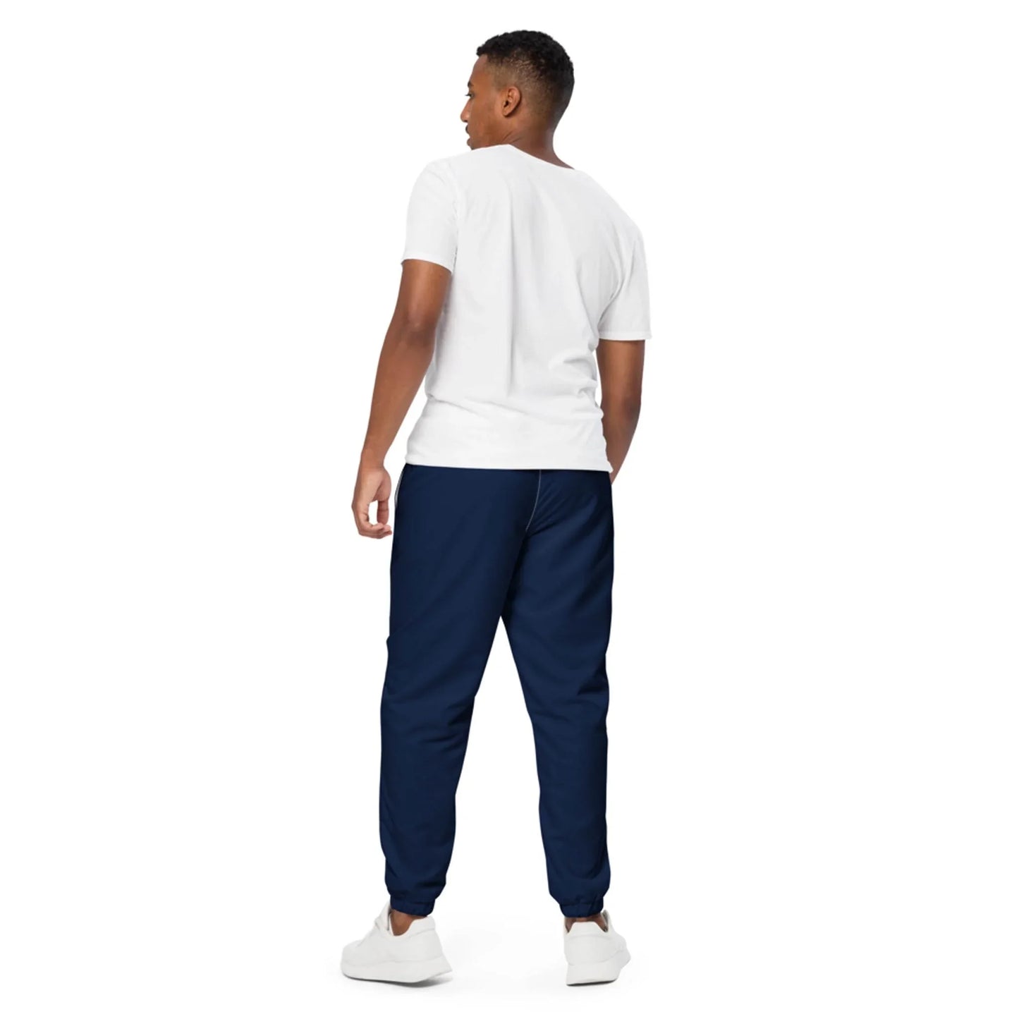 A man stands facing away from the camera, wearing a Mambí white t-shirt, navy blue jogger pants, and white sneakers against a plain white background.