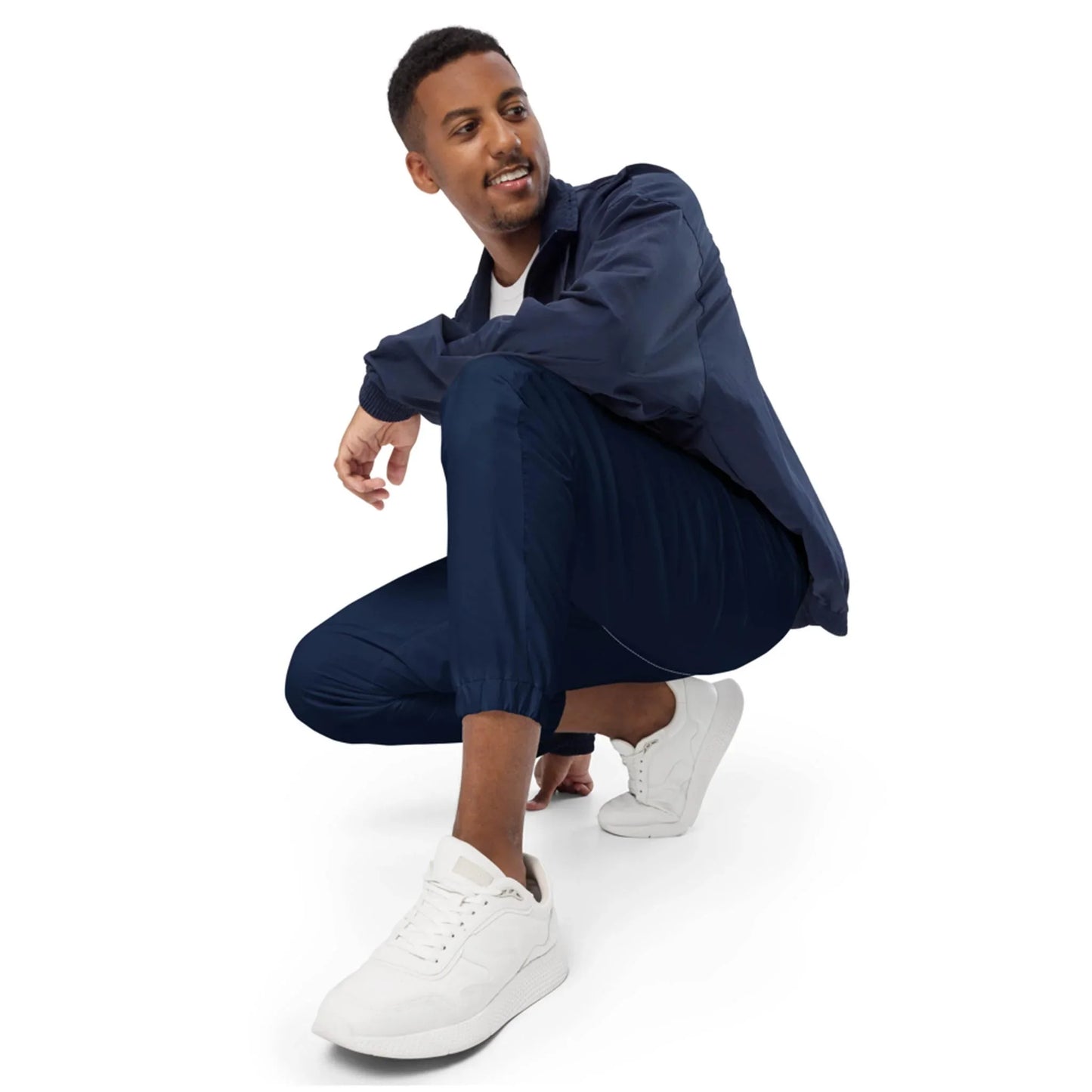 A man wearing a navy blue Mambí jacket and pants with white sneakers is squatting and smiling, looking off to the side against a plain white background.