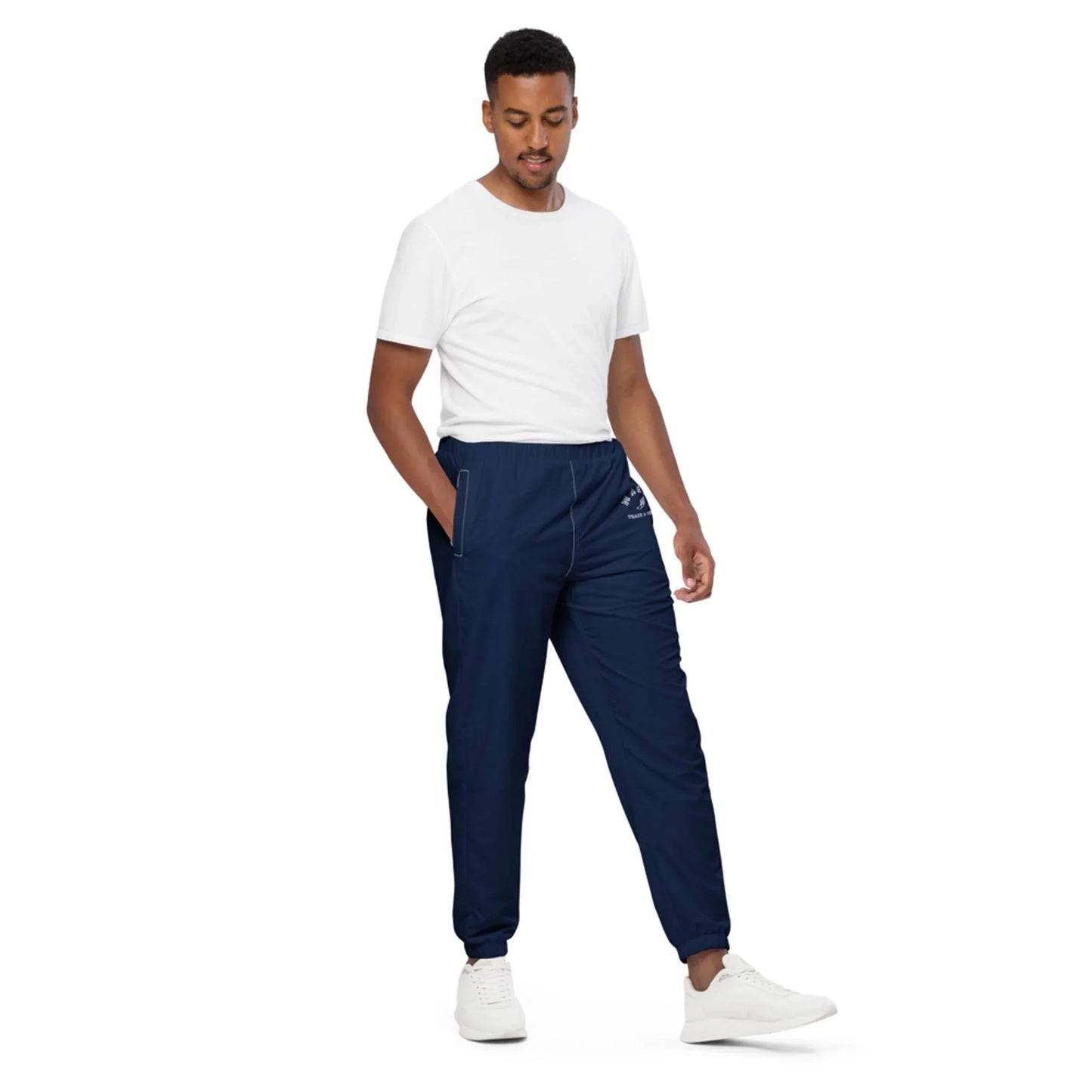 A man wearing a plain white t-shirt, navy blue Mambí jogger pants with a small logo, and white sneakers stands with one hand in his pocket against a white background.