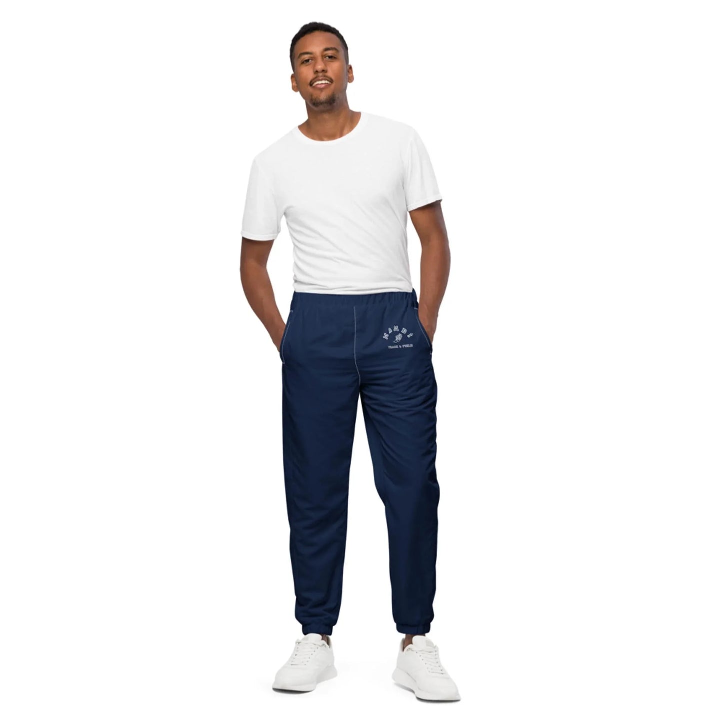 A man stands against a white background wearing a plain white t-shirt, navy blue Mambí jogger pants with a small logo on the hip, and white sneakers. His hands are in his pockets as he looks at the camera.