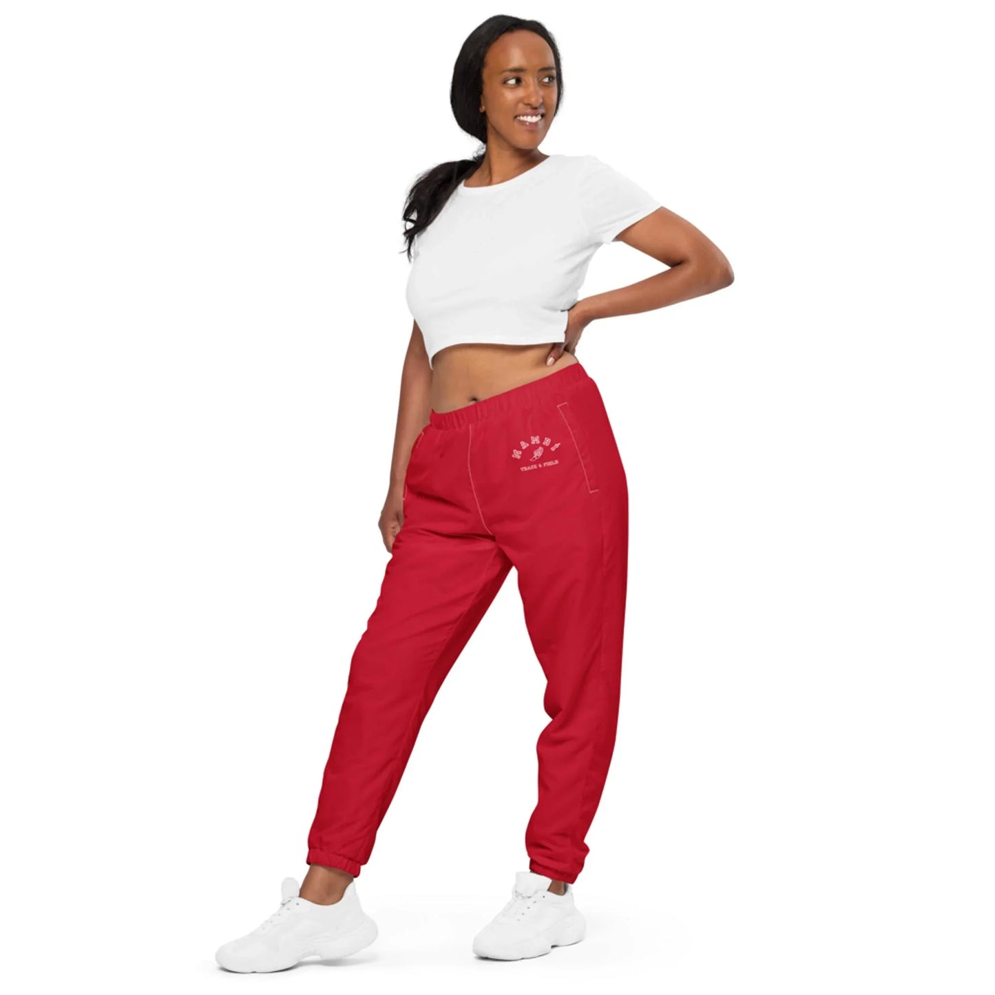 A woman in a white Mambí crop top and red jogger pants stands sideways, looking to her left and smiling. She poses with one hand on her hip, wearing white sneakers against a plain white background.