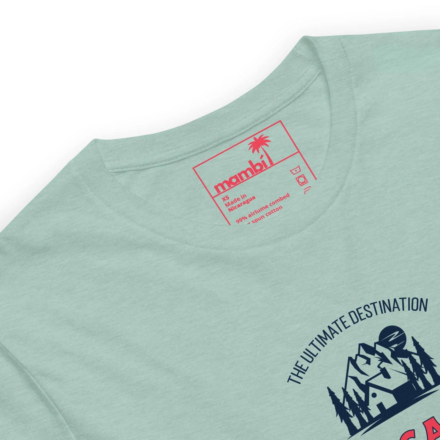 Close-up of a pale green Mambí t-shirt with a red label printed inside the collar, showing size and care instructions. Part of a mountain and house graphic with the text “THE ULTIMATE DESTINATION” is visible on the shirt front.
