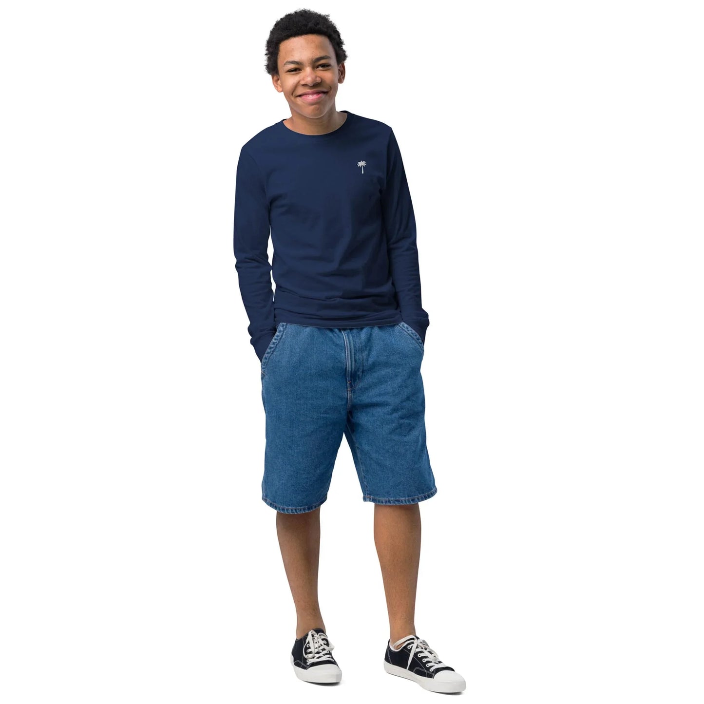 A young person stands smiling with hands in the pockets of blue denim shorts, wearing a navy Mambí long-sleeve shirt and black sneakers with white soles, against a white background.