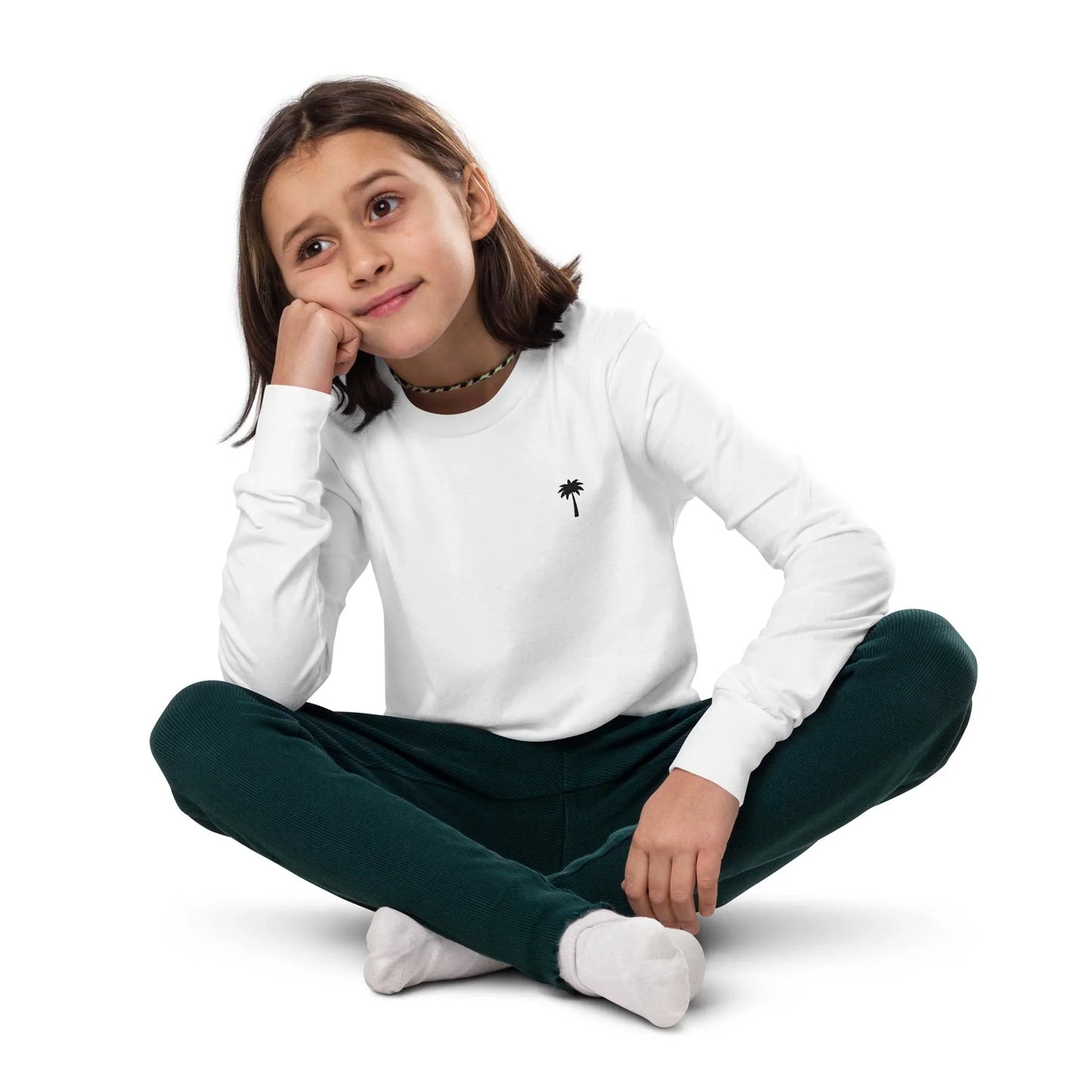A young child with shoulder-length brown hair sits cross-legged on the floor, wearing a white Mambí long-sleeve shirt with a small palm tree logo, dark green pants, and white socks, looking thoughtful with their head resting on one hand.