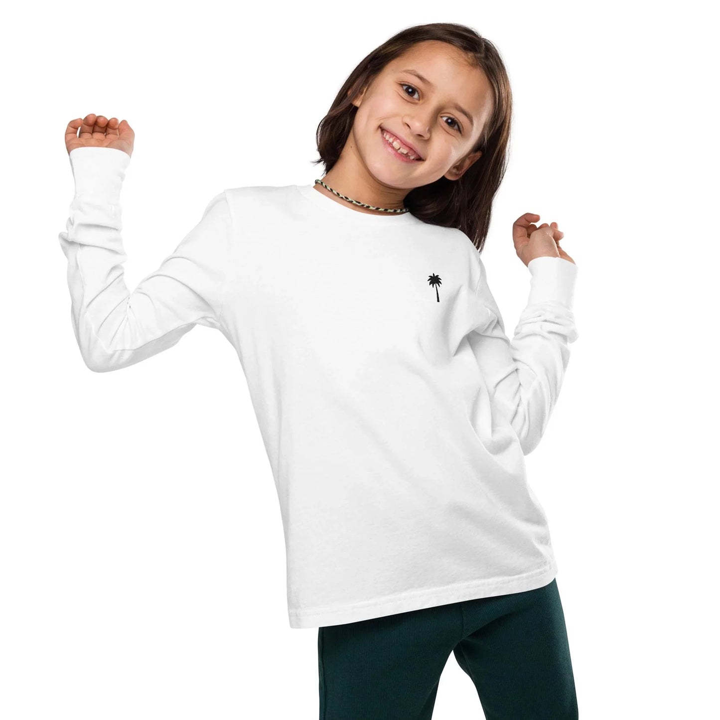 A smiling child with shoulder-length brown hair wears a white long-sleeve Mambí shirt featuring a small black palm tree graphic and dark pants, striking a playful pose against a plain white background.