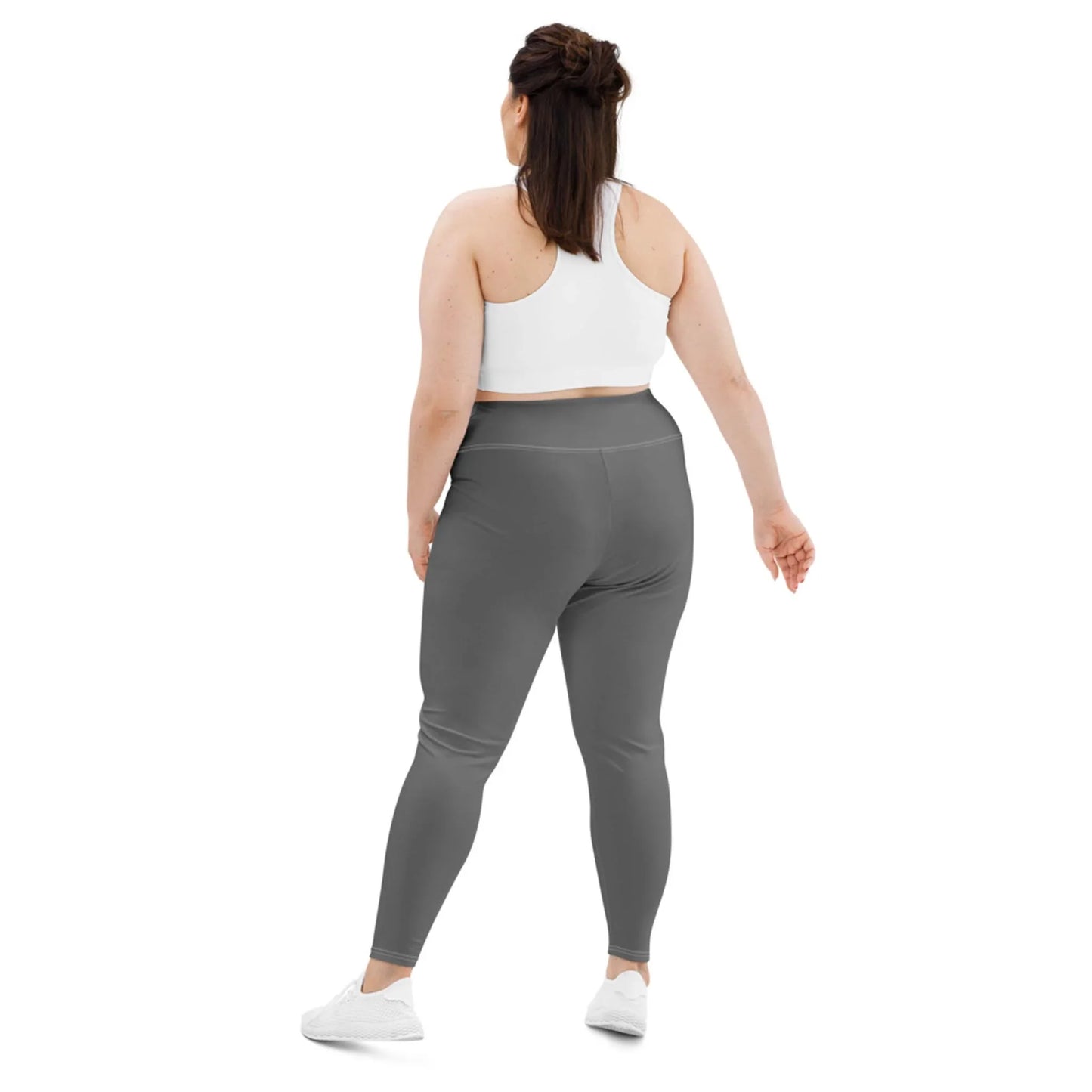 A woman with long dark hair wears a Mambí white sports bra, gray high-waisted leggings, and white sneakers. She stands facing away, body slightly turned to the side, against a plain white background.
