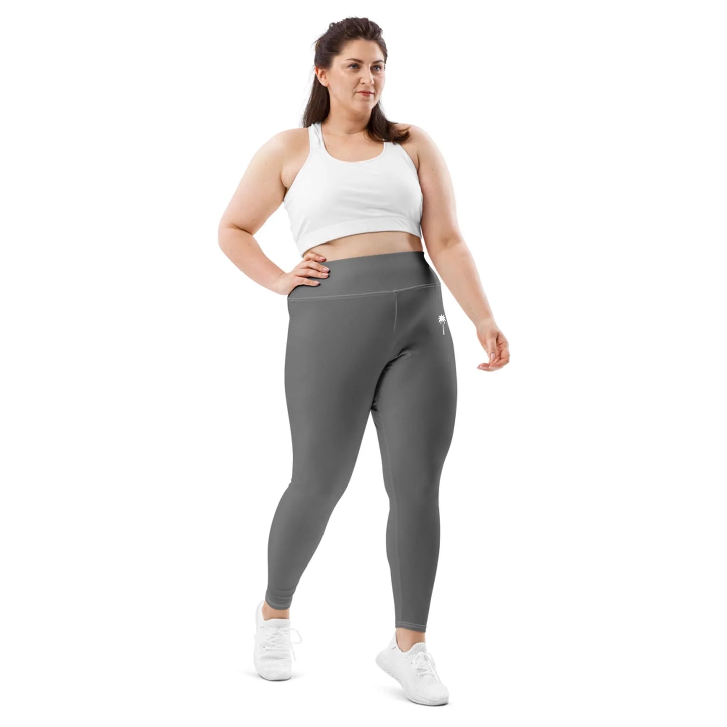 A woman wearing a Mambí white sports bra, gray high-waisted leggings, and white sneakers stands against a plain white background.