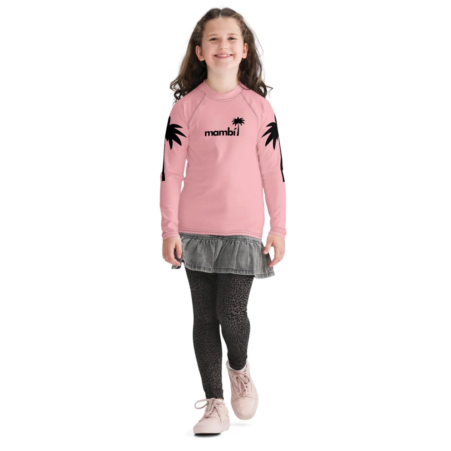 A smiling young girl with long brown hair wears a pink long-sleeve shirt featuring Mambí text and black palm tree graphics, a gray skirt, patterned leggings, and pink shoes as she walks forward.