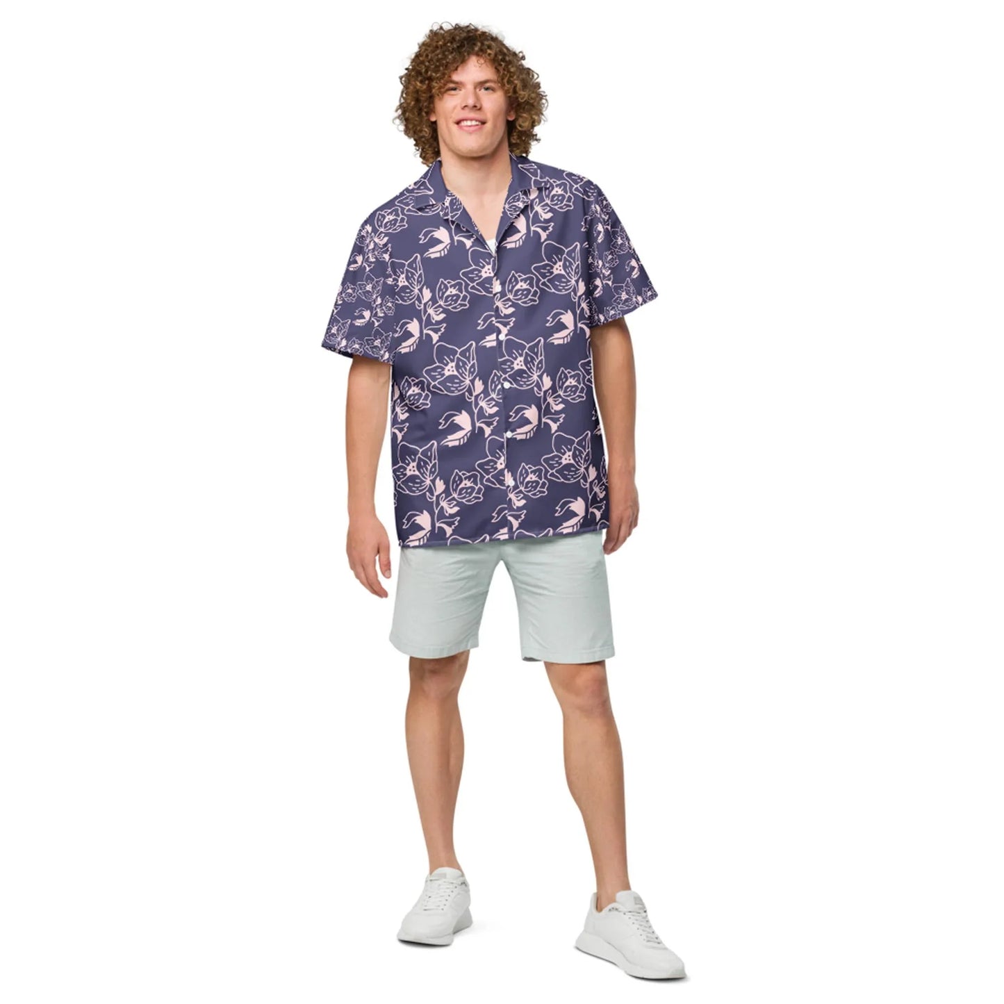 A man with curly hair wearing a Mambí purple floral short-sleeve shirt, light gray shorts, and white sneakers stands smiling against a white background.