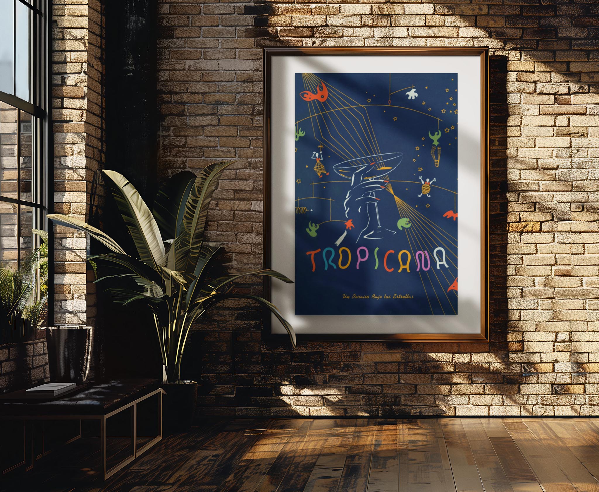 Framed “Tropicana” art poster hangs on a brick wall in a sunlit room with wood floors, a large plant, and tall windows; sunlight casts shadows across the wall and floor.