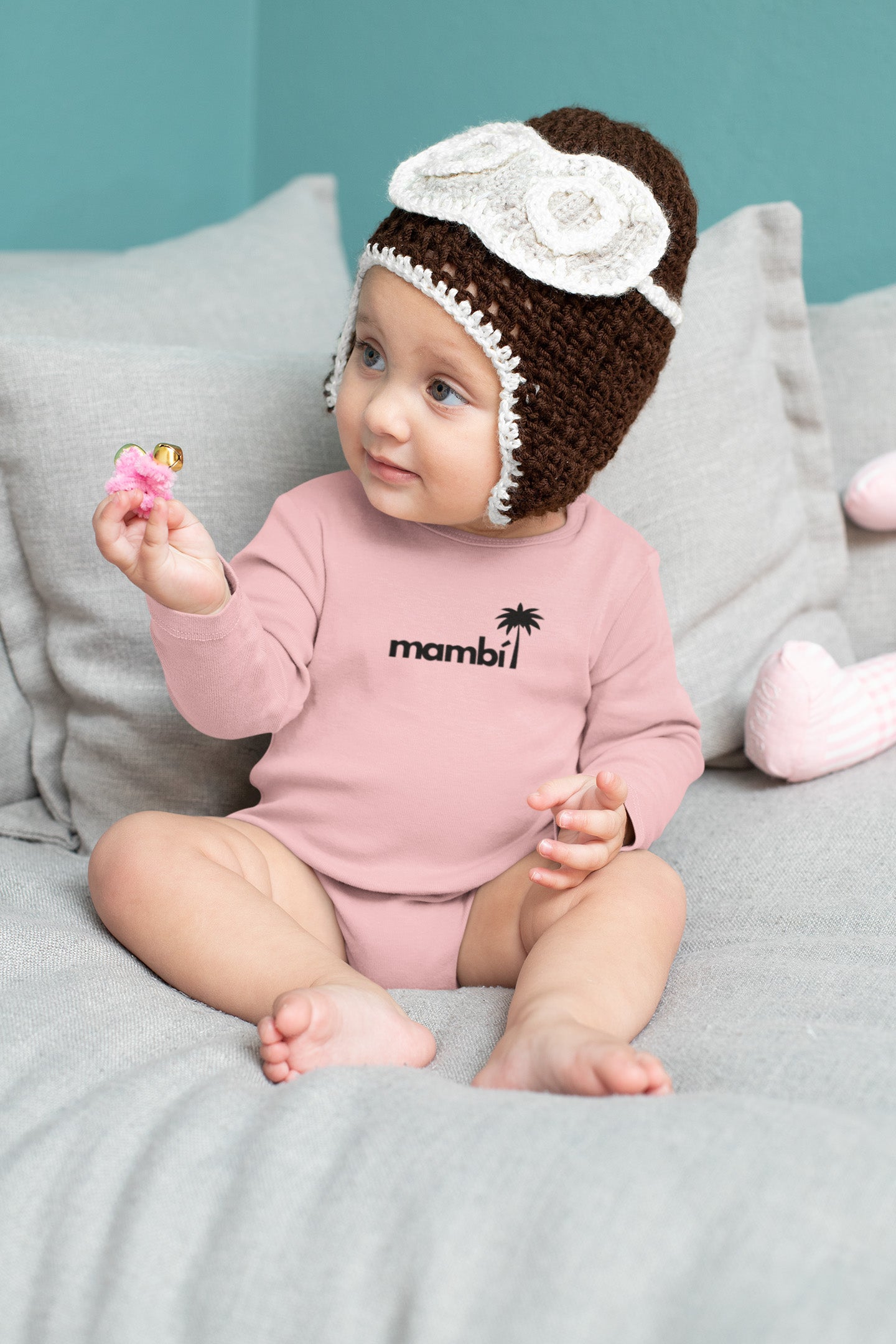 A baby wearing a crocheted aviator hat and a pink onesie with the word mambi and a palm tree logo sits on a gray couch, holding a small pink toy and looking to the side.