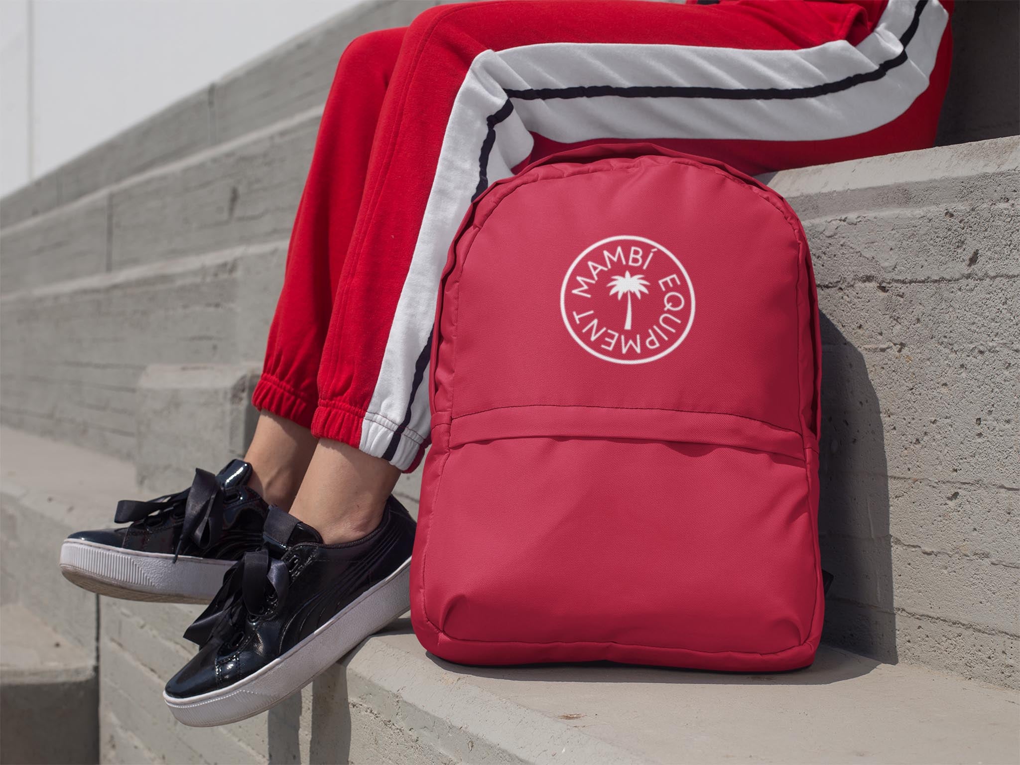 Person wearing red track pants with a white and black stripe sits on concrete steps next to a matching red backpack with a white circular logo that reads AMBIENTE EQUIPMENT. Black sneakers with black bows are visible.