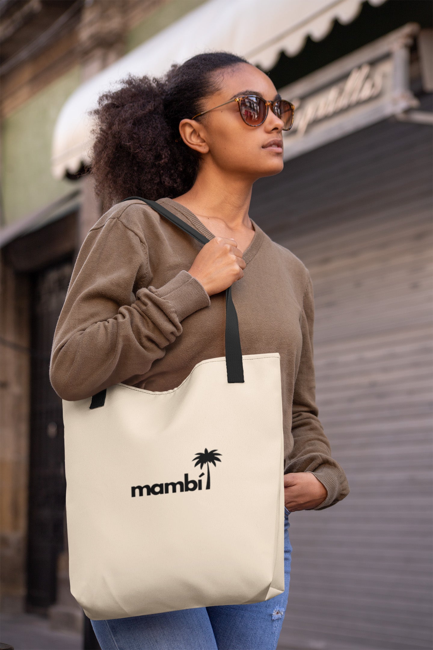 A woman wearing sunglasses and a brown sweater holds a cream-colored tote bag with black straps. The bag has the word mambi and a palm tree graphic printed on it. She stands outdoors in front of a closed shop.