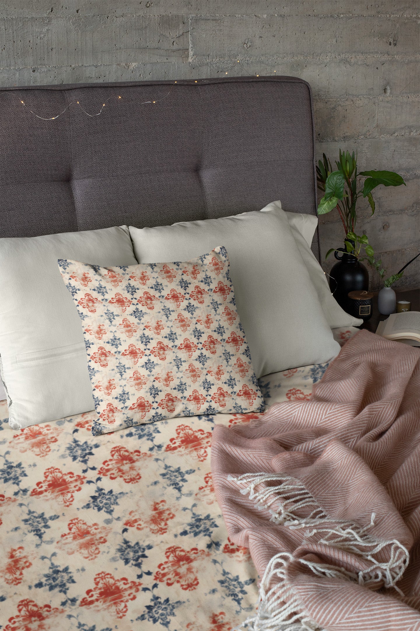 A neatly made bed with light gray pillows, a floral-patterned pillow, a matching floral bedspread, and a pinkish striped throw blanket. A plant, lamp, and book sit on the bedside table against a concrete wall.