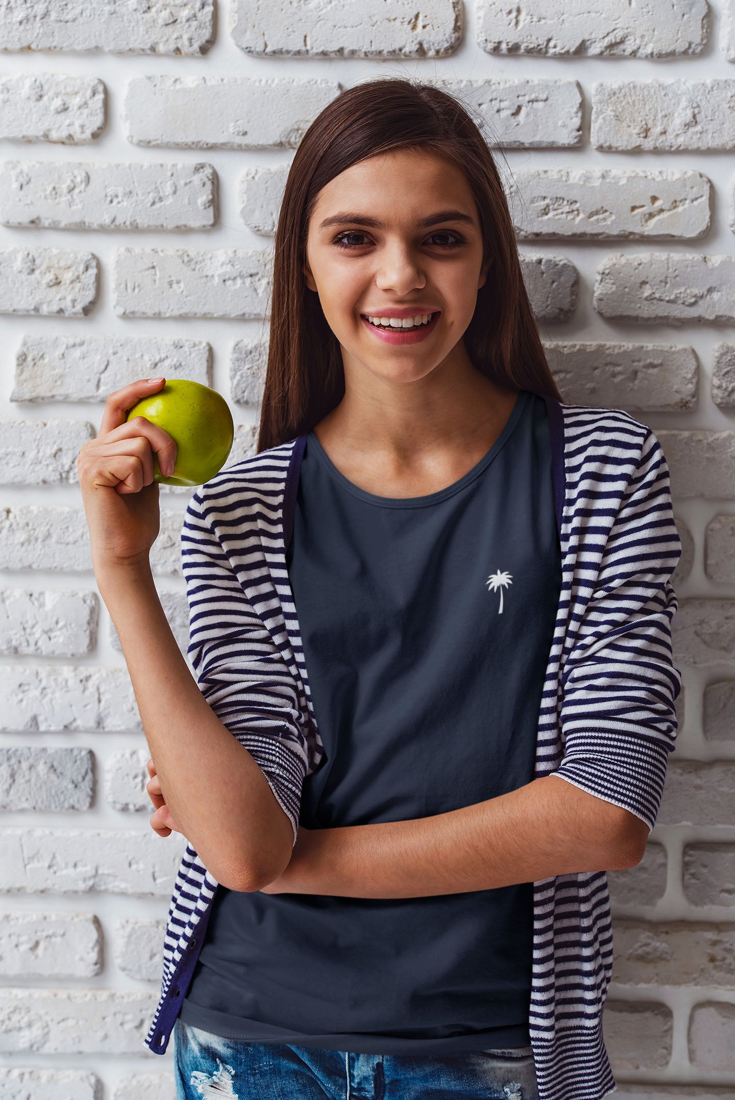 A smiling teenage girl with long brown hair, wearing a navy striped cardigan and holding a green apple, stands in front of a white brick wall.