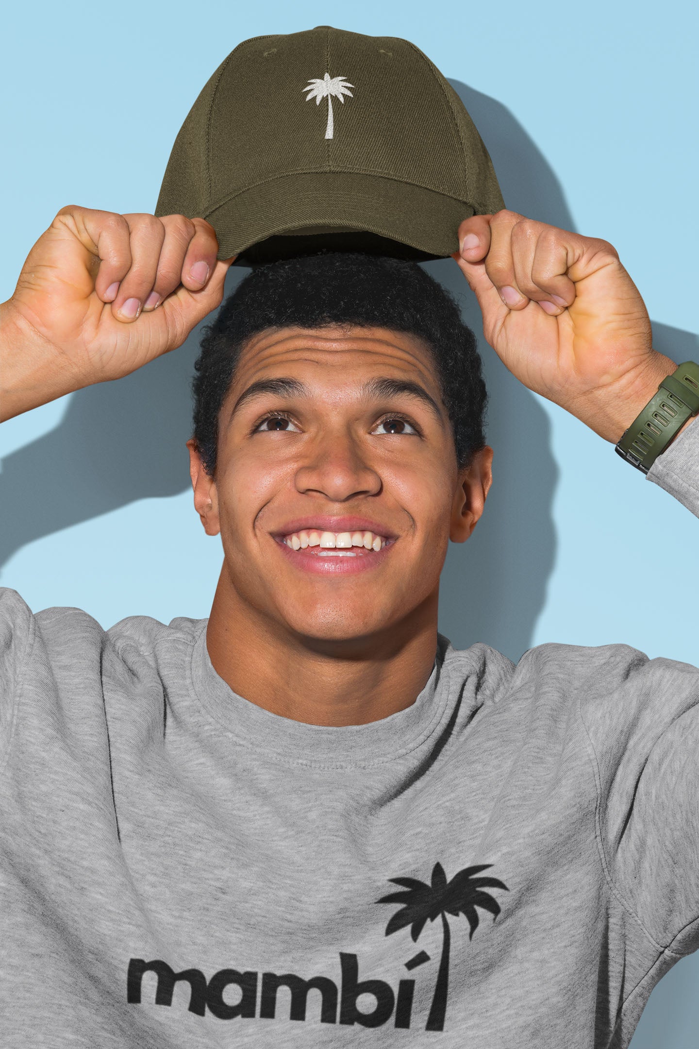 A smiling young man in a gray mambi sweatshirt lifts an olive green cap with a palm tree logo above his head, posing against a light blue background.
