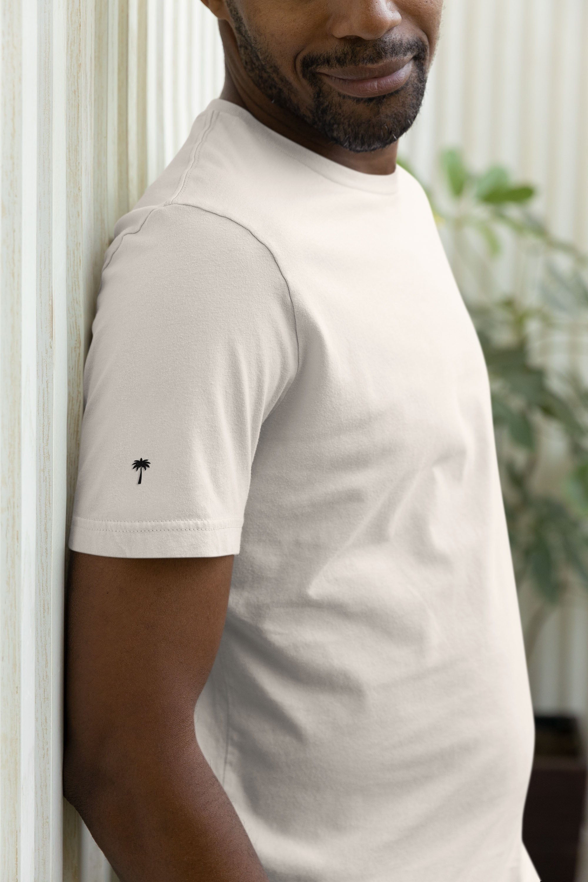 A man in a light beige t-shirt with a small black palm tree graphic on the sleeve leans against a wall, smiling slightly. A green plant is visible in the blurred background.
