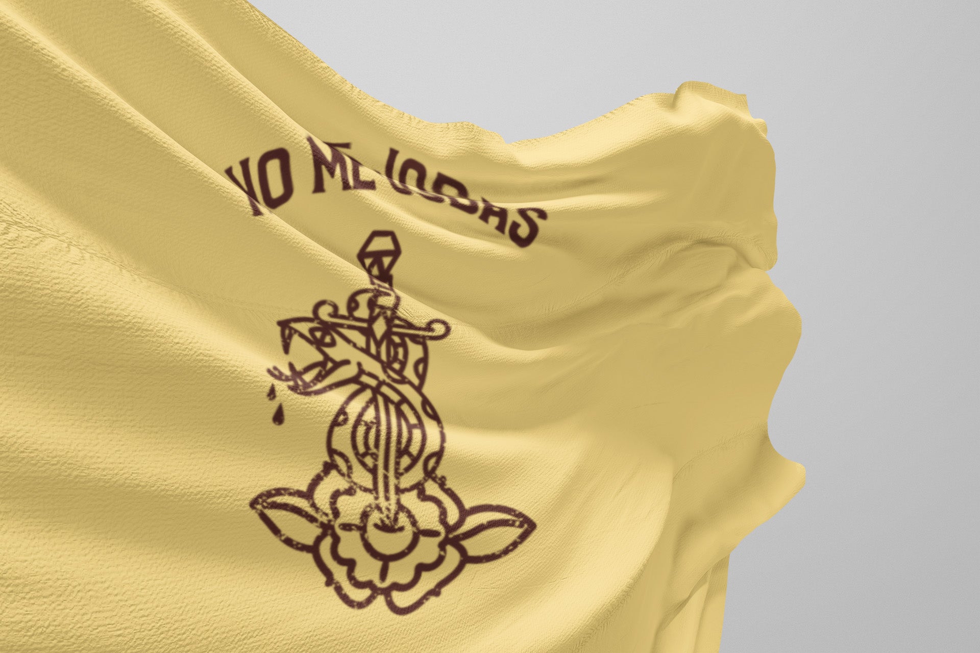 A yellow fabric with a textured surface features a dark illustration of a traditional-style dagger and flower, along with the phrase NO ME LLORES in bold, curved letters above the design.