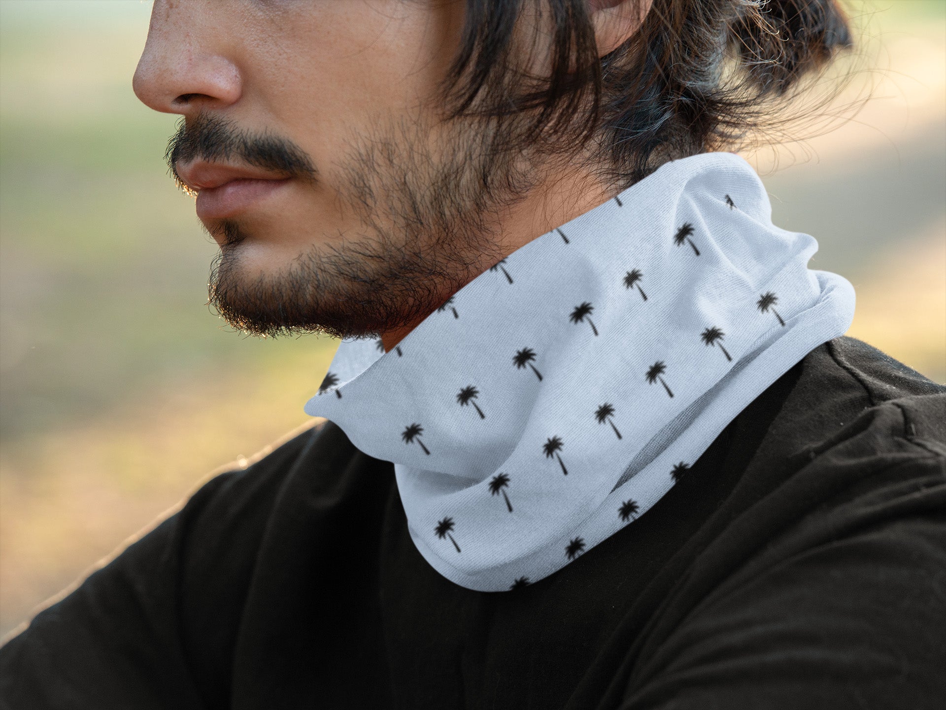 A person with a short beard and mustache wears a white neck gaiter patterned with small black palm trees, paired with a black shirt, outdoors in soft natural light.