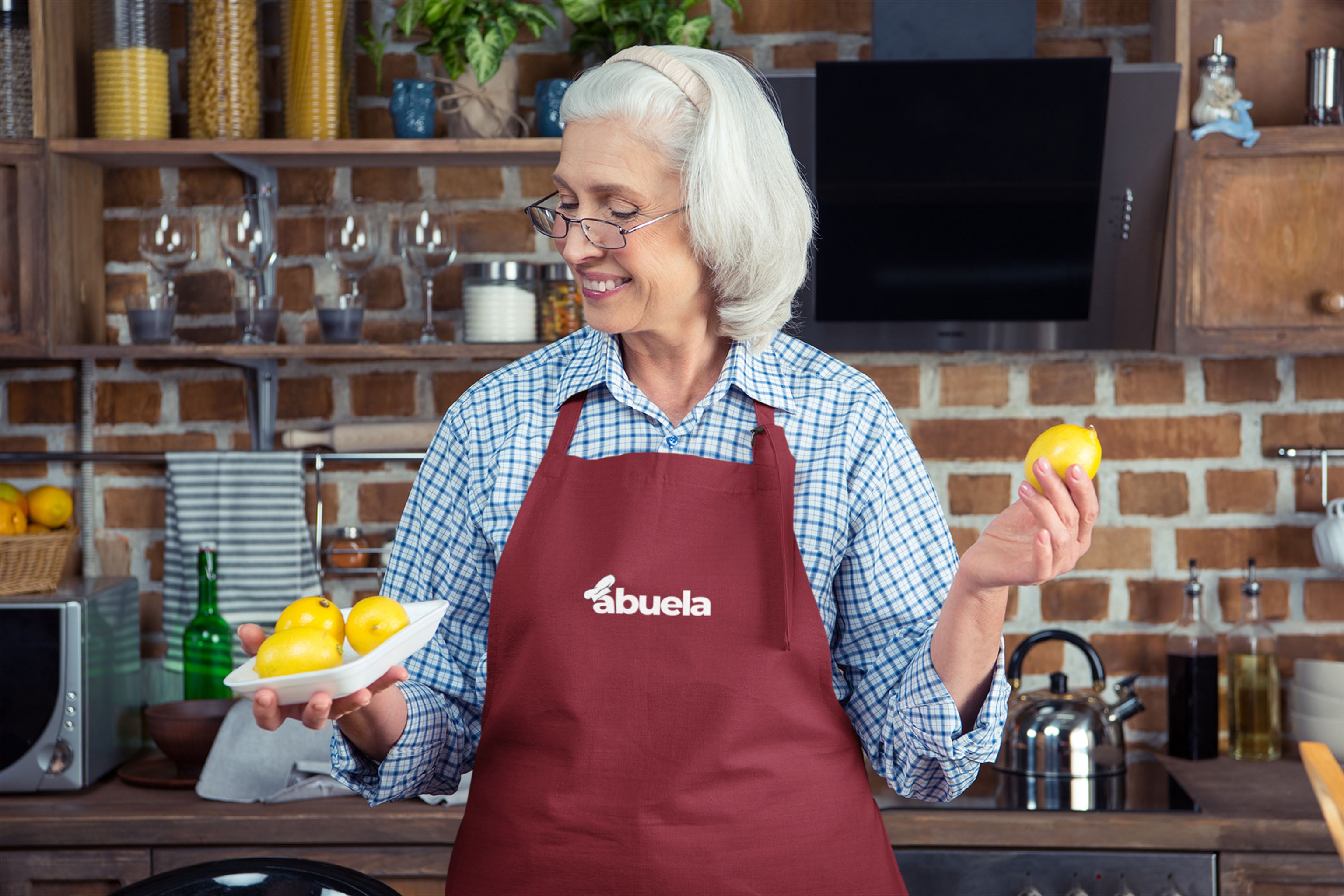 An older woman with gray hair and glasses, wearing a red apron that says abuela, holds a plate of lemons in one hand and a lemon in the other, standing in a cozy kitchen with a brick wall and wooden shelves.