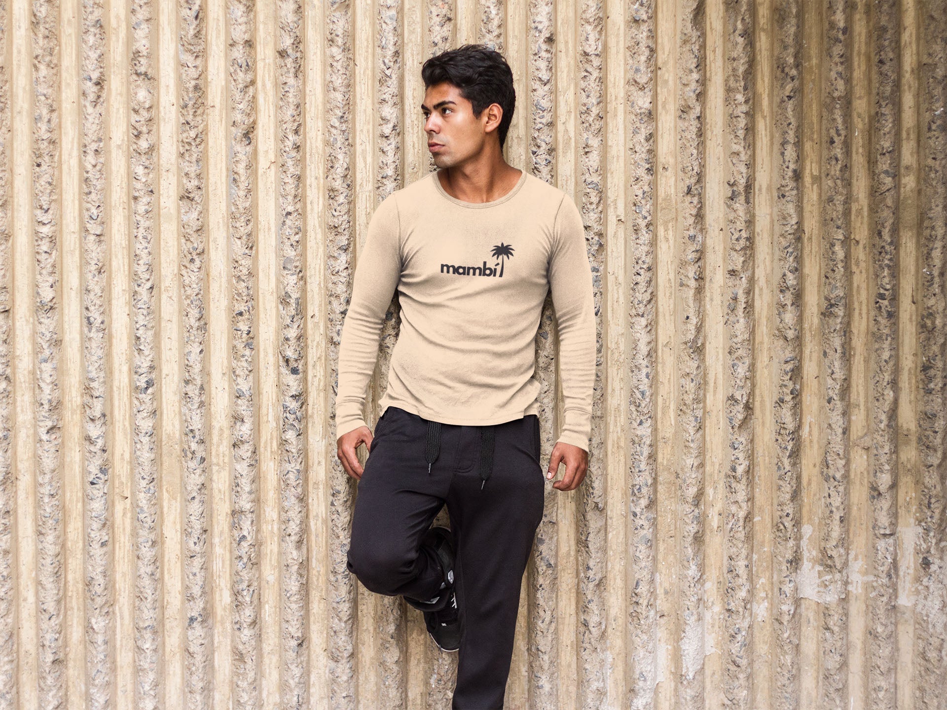 A man wearing a beige long-sleeve shirt with the word mambi and a palm tree graphic stands against a textured, vertical, corrugated wall with one foot resting on the wall and hands in his pockets.