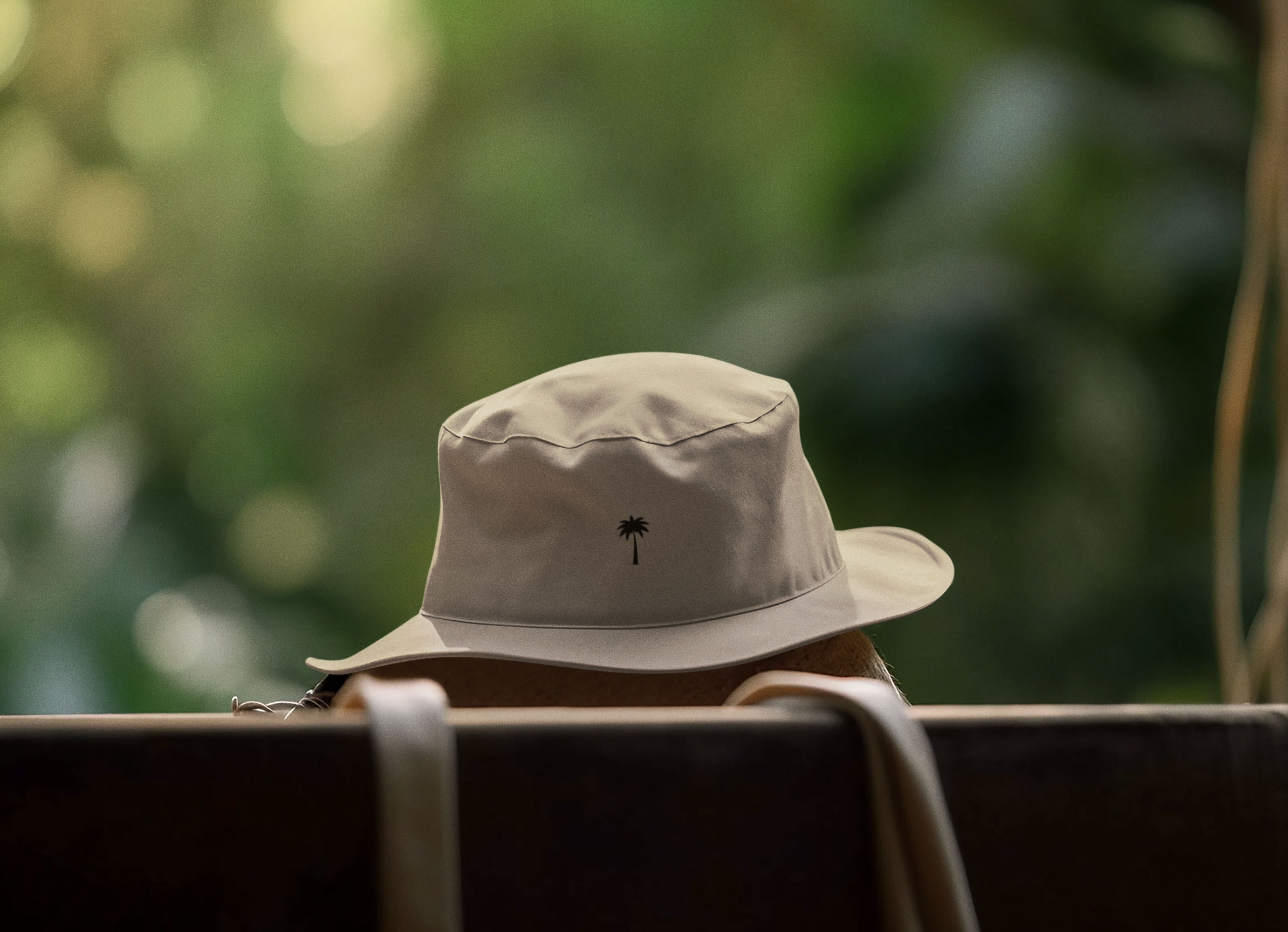 A light-colored bucket hat with a small palm tree design sits on a surface, with a blurred green outdoor background. Straps or handles hang in the foreground.