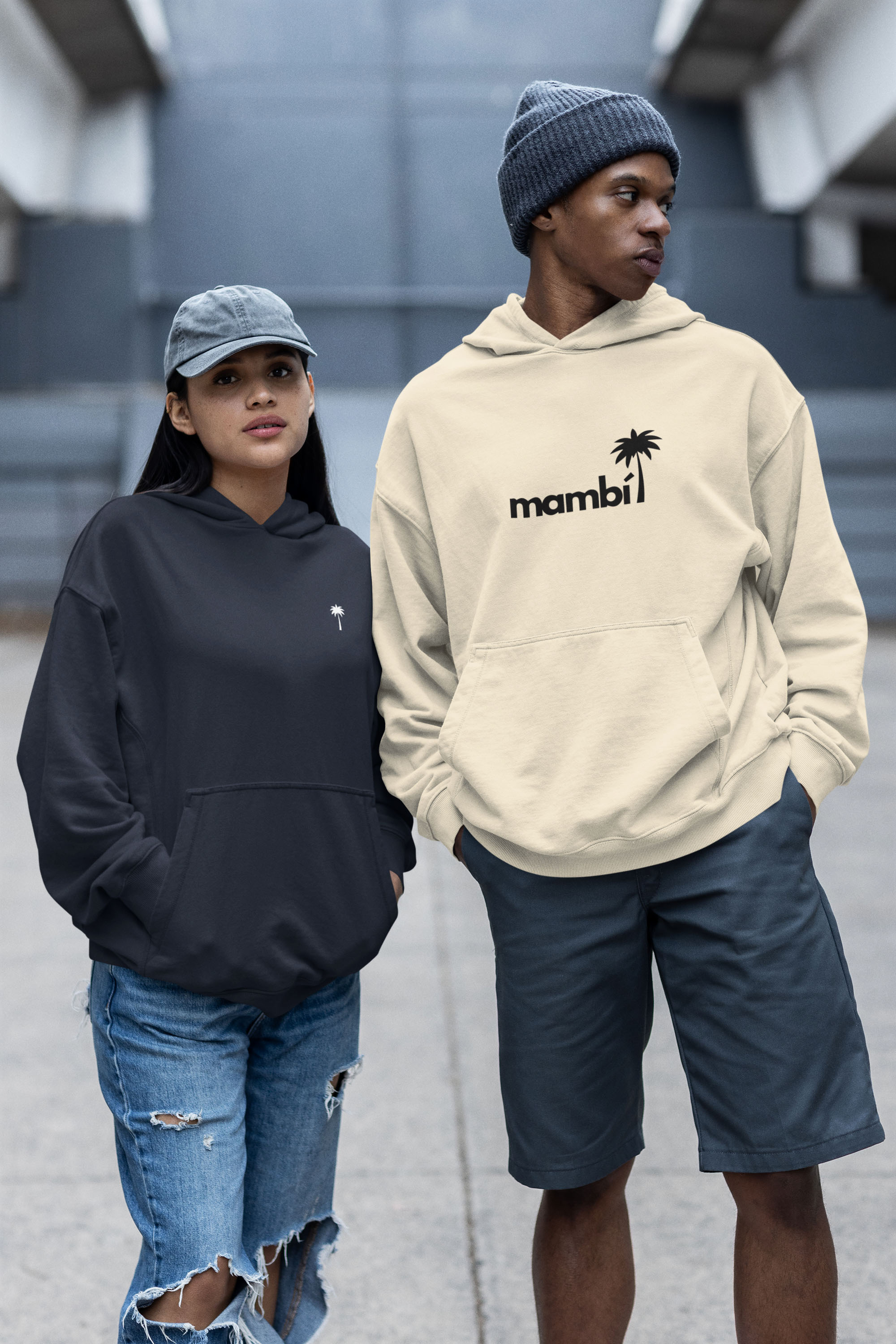Two people stand side by side outdoors wearing hoodies and hats. One wears a black hoodie and ripped jeans; the other wears a beige hoodie with mambi and a palm tree graphic, gray shorts, and a knit beanie.