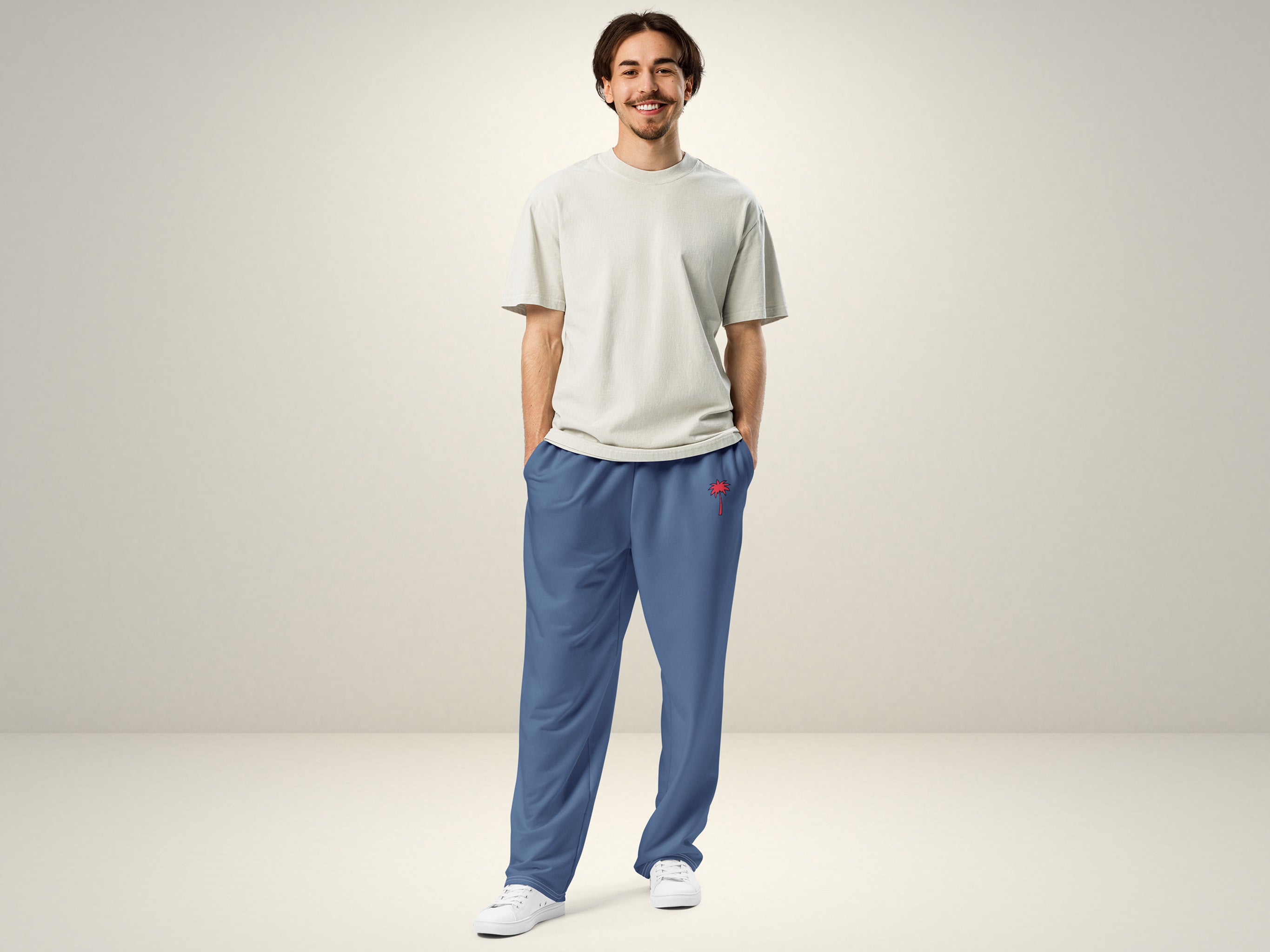 A smiling man stands with hands in the pockets of his blue pants, wearing a light grey t-shirt and white sneakers, against a plain light background.