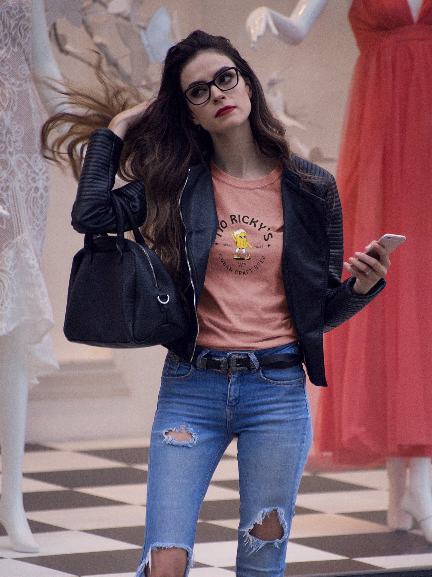 A woman with long brown hair and glasses flips her hair while holding a phone and a black handbag. She wears a black jacket, pink T-shirt, and ripped jeans. Mannequins with dresses are seen in the background.
