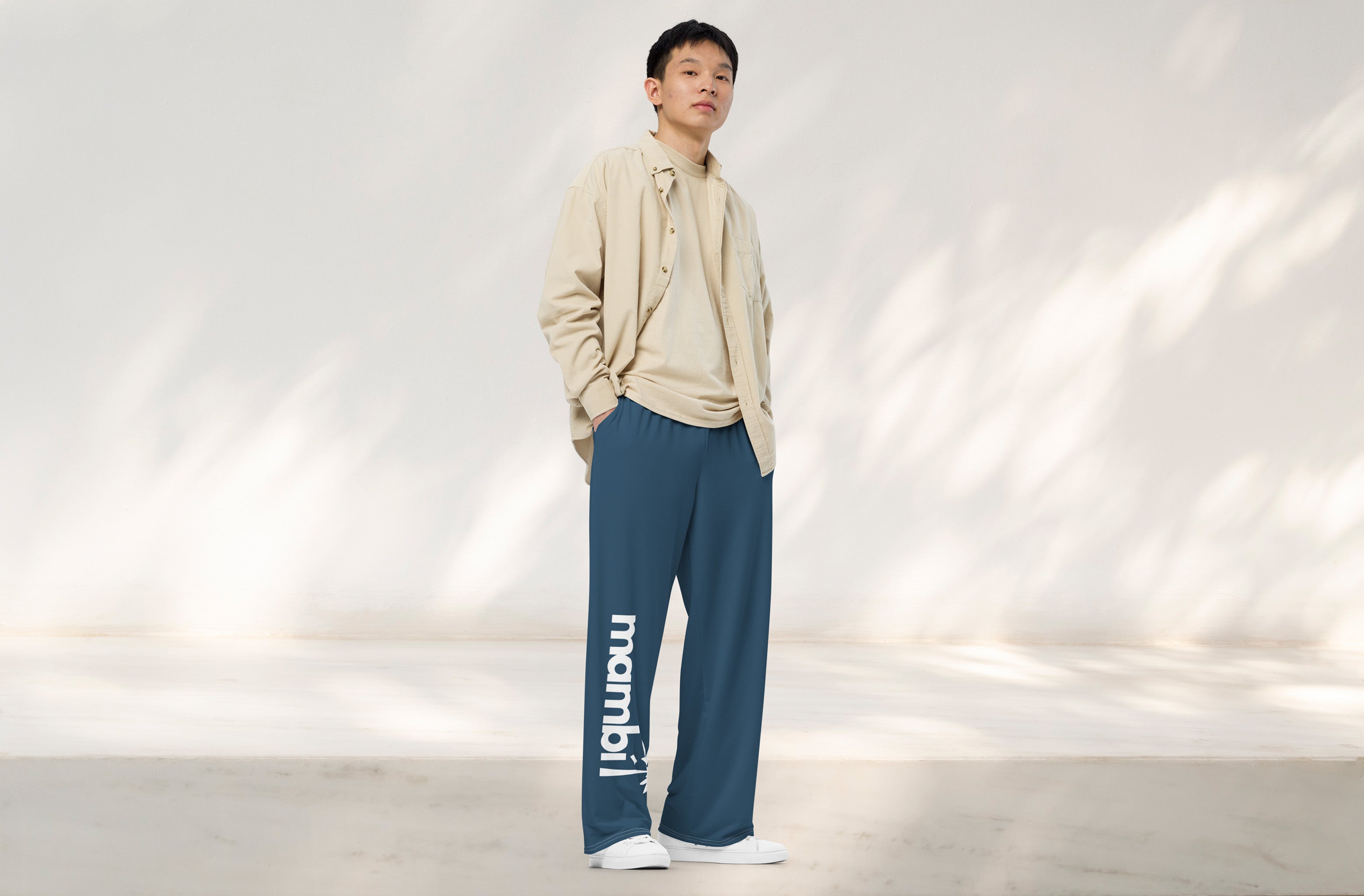 A person stands indoors against a light wall, wearing a beige shirt and t-shirt, blue wide-leg pants with “mambi” written vertically in white on one leg, and white sneakers.