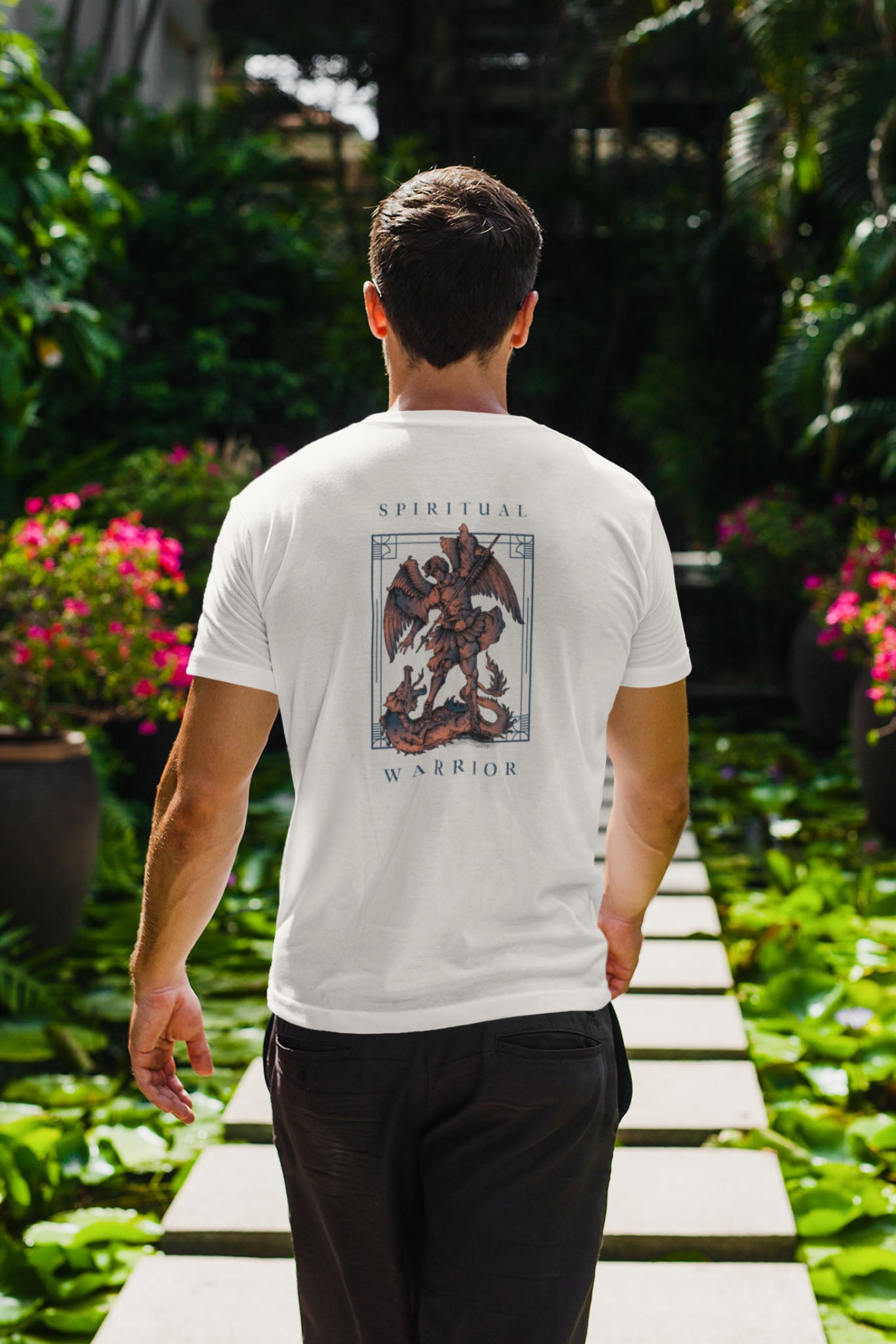 A man walks on a stone path through a garden with lush greenery and pink flowers, wearing a white T-shirt with an illustrated angel and the words Spiritual Warrior on the back.