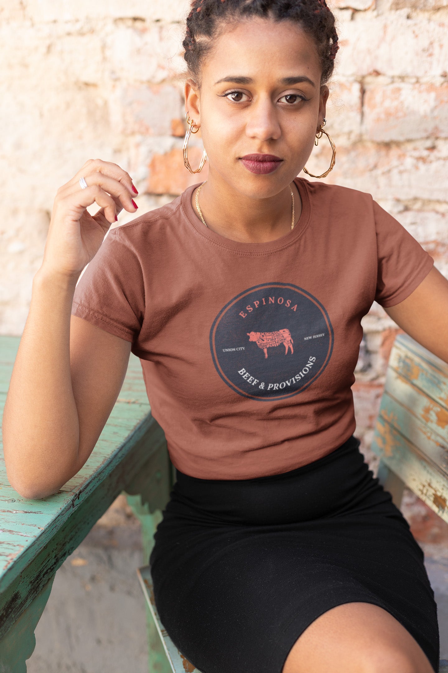 A woman with braided hair sits at a weathered green table, wearing a brown t-shirt with a circular logo that reads Espinosa Beef & Provisions and a black skirt, in front of a rustic brick wall.